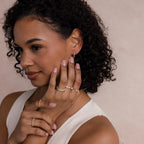 A woman with curly hair wears gold rings and Pave Pearl Huggies earrings, posing with her hands near her face against a neutral background.