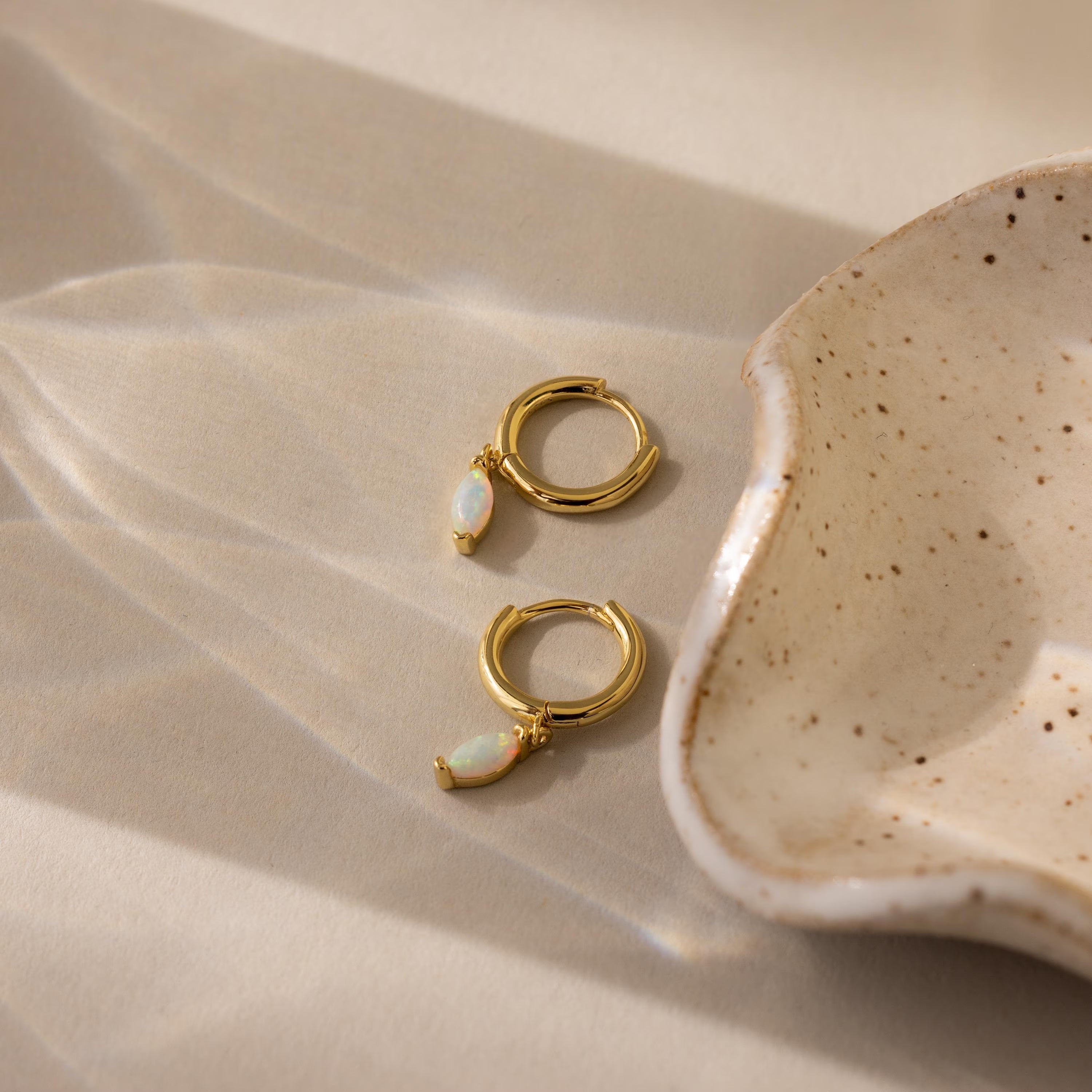 Opal Marquise Huggies featuring small opal charms are displayed next to a speckled ceramic dish on a beige surface, bringing an elegant touch to any setting.