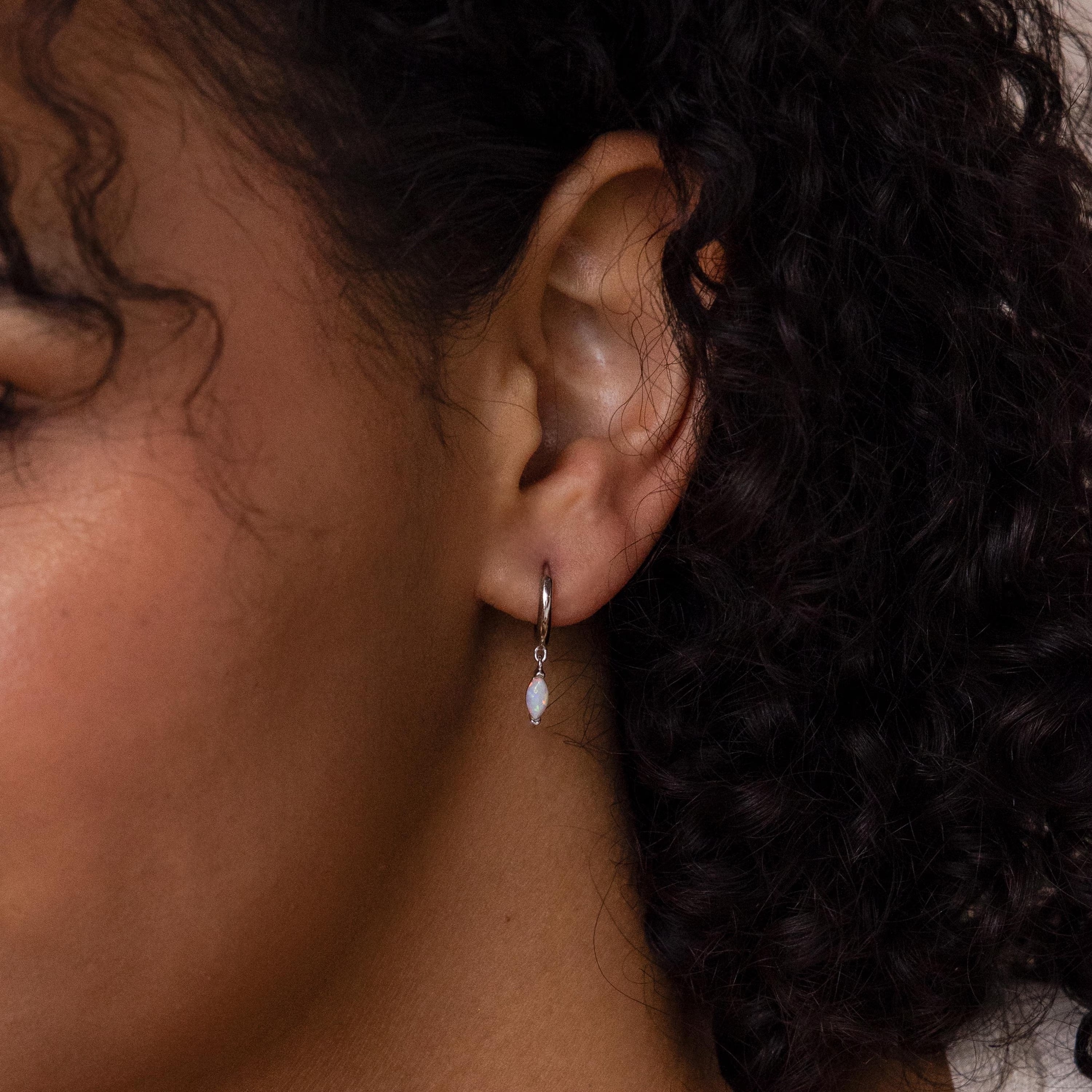 Close-up of a person with curly hair wearing Opal Marquise Huggies—delicate hoops featuring a small hanging opal gemstone.