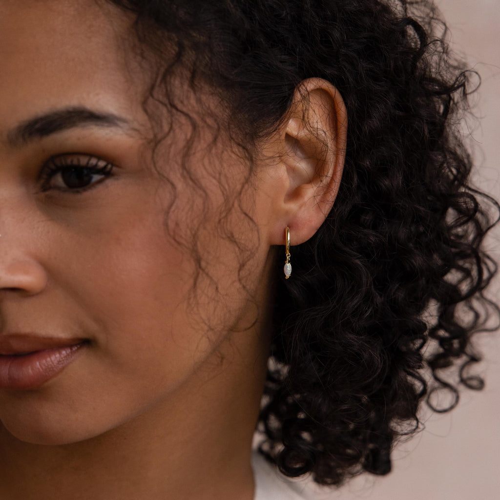 A woman with curly hair wears the Opal Marquise Huggies—small gold hoop earrings featuring a dangling marquise-cut opal charm.