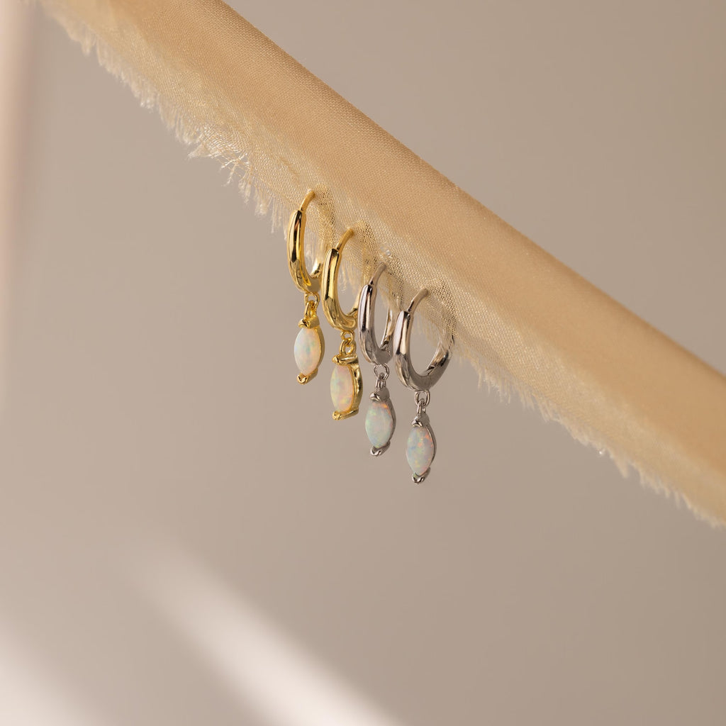 Two pairs of Opal Marquise Huggies, one in gold and one in silver, displayed on a beige textured fabric strip.