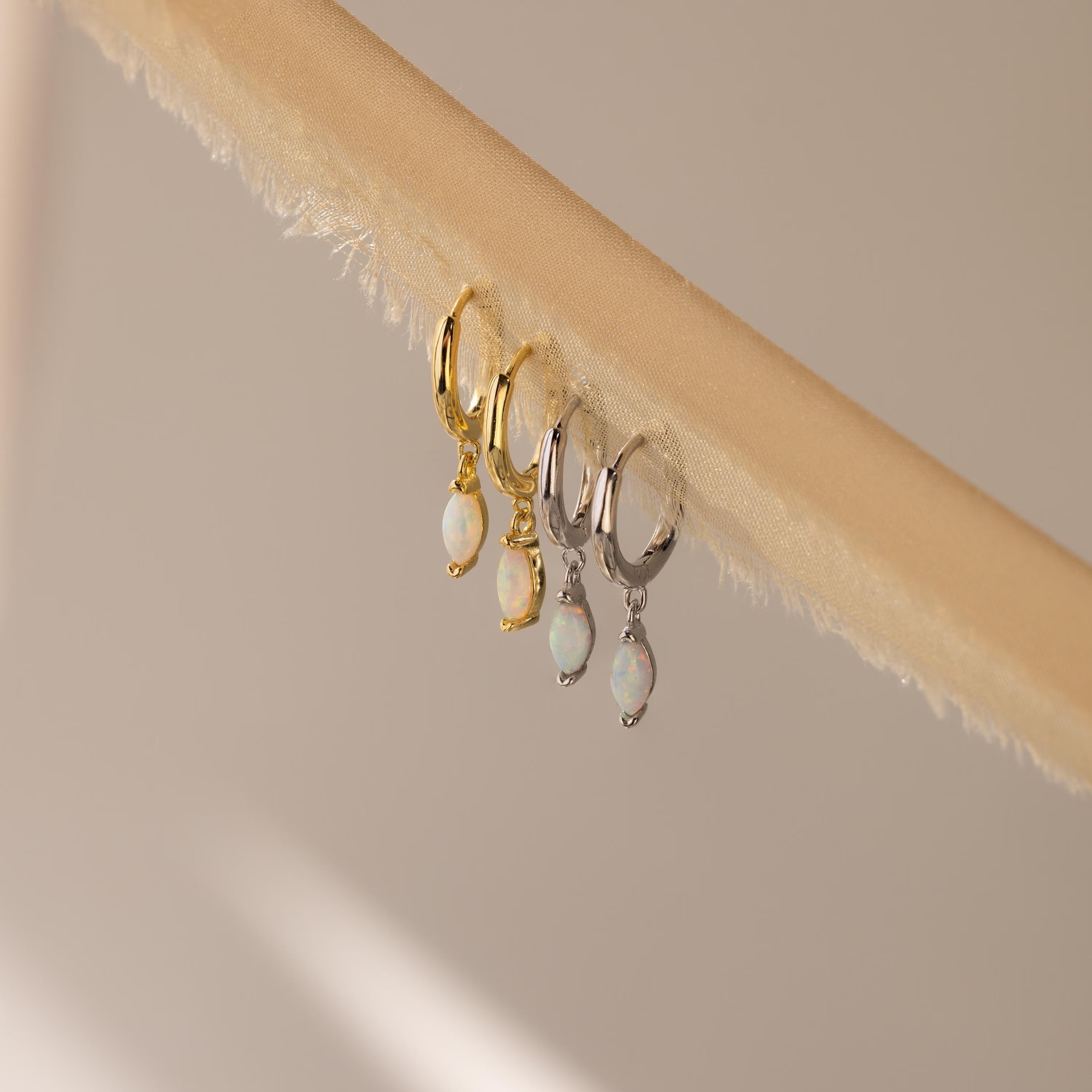 Two pairs of Opal Marquise Huggies, one in gold and one in silver, displayed on a beige textured fabric strip.