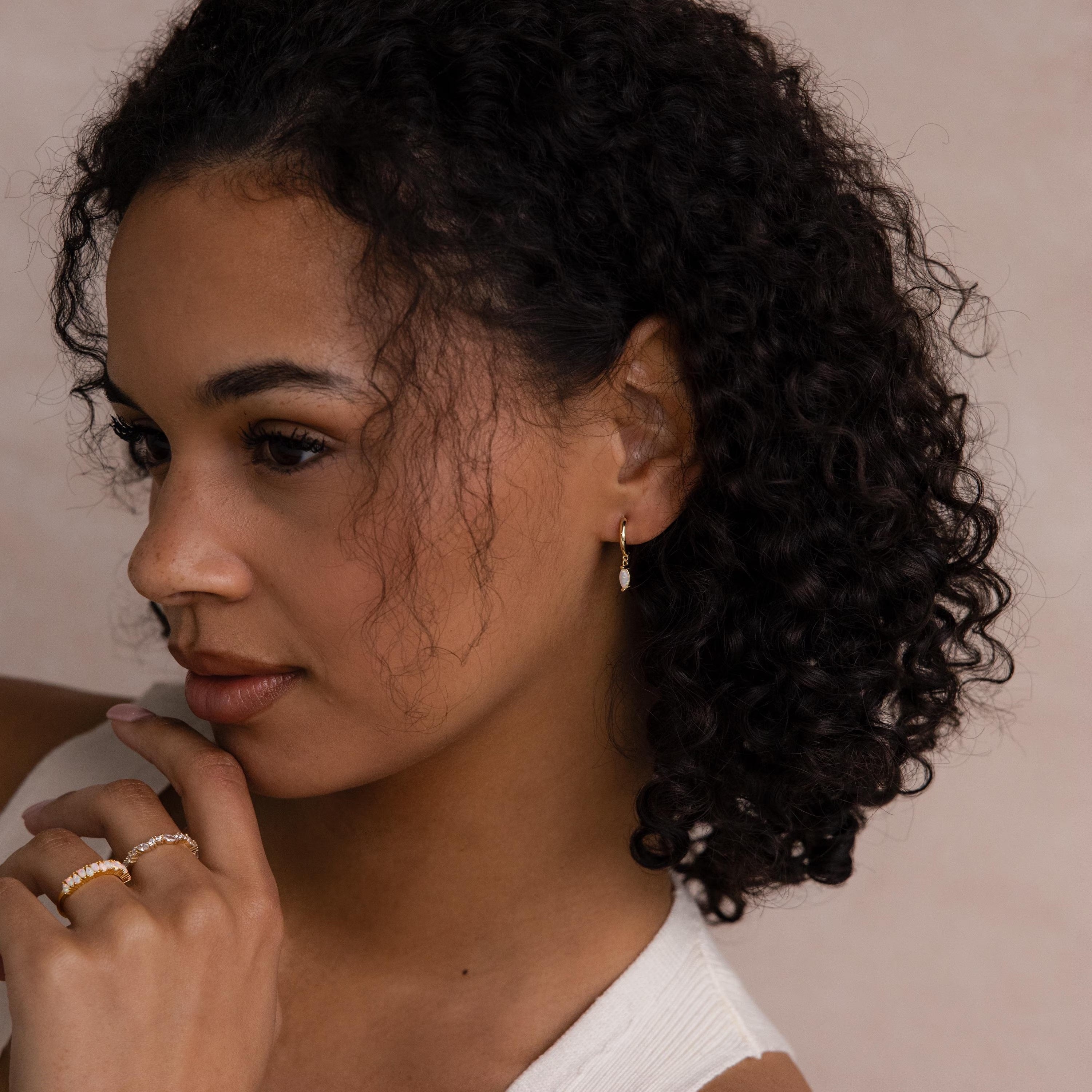 Woman with curly hair wearing Opal Marquise Huggies and rings, touching her chin and looking to the side.