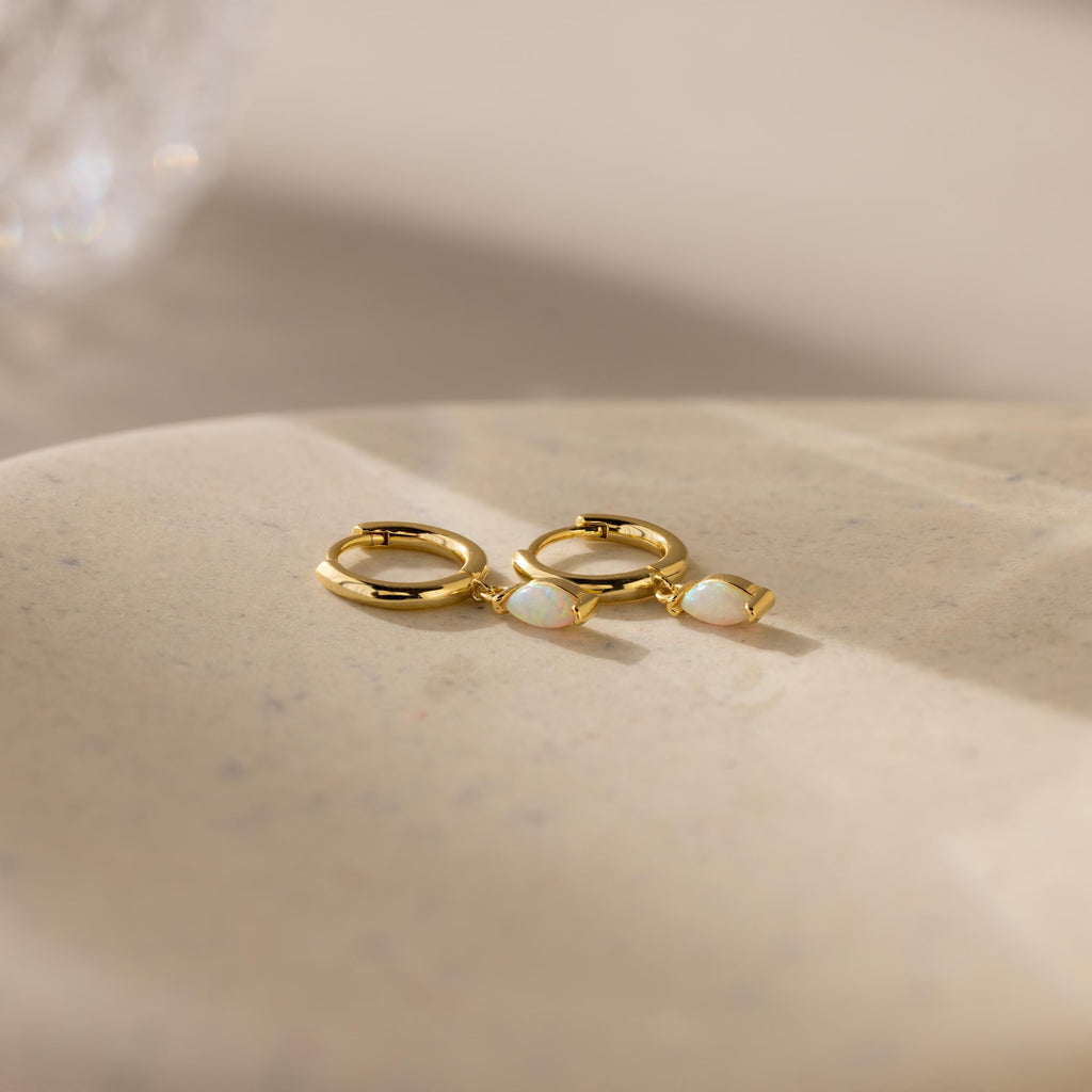 The Opal Marquise Huggies, featuring two gold hoops with white opal stones, rest on a smooth beige surface in soft lighting—the perfect pair for understated elegance.