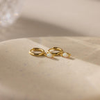 The Opal Marquise Huggies, featuring two gold hoops with white opal stones, rest on a smooth beige surface in soft lighting—the perfect pair for understated elegance.