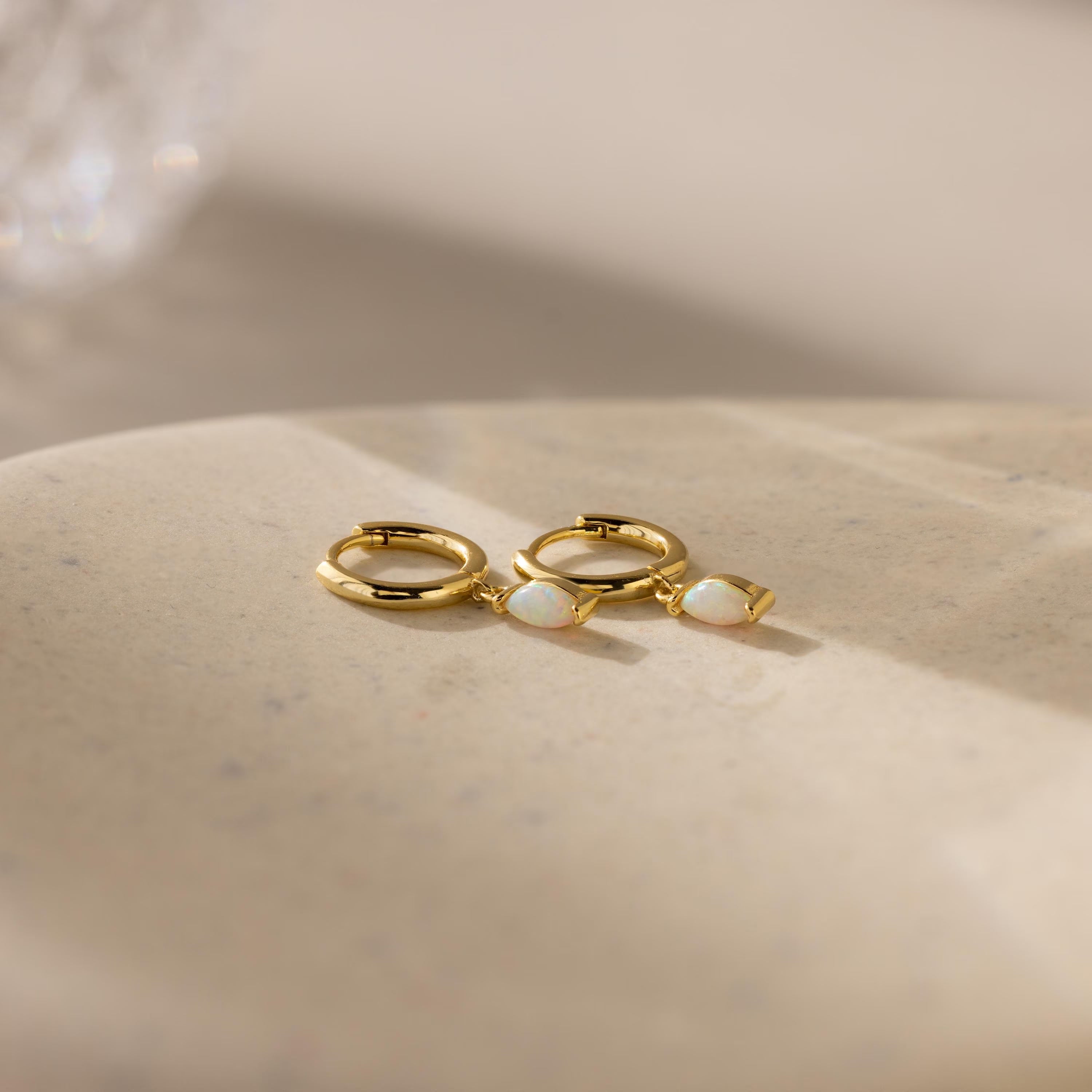 The Opal Marquise Huggies, featuring two gold hoops with white opal stones, rest on a smooth beige surface in soft lighting—the perfect pair for understated elegance.