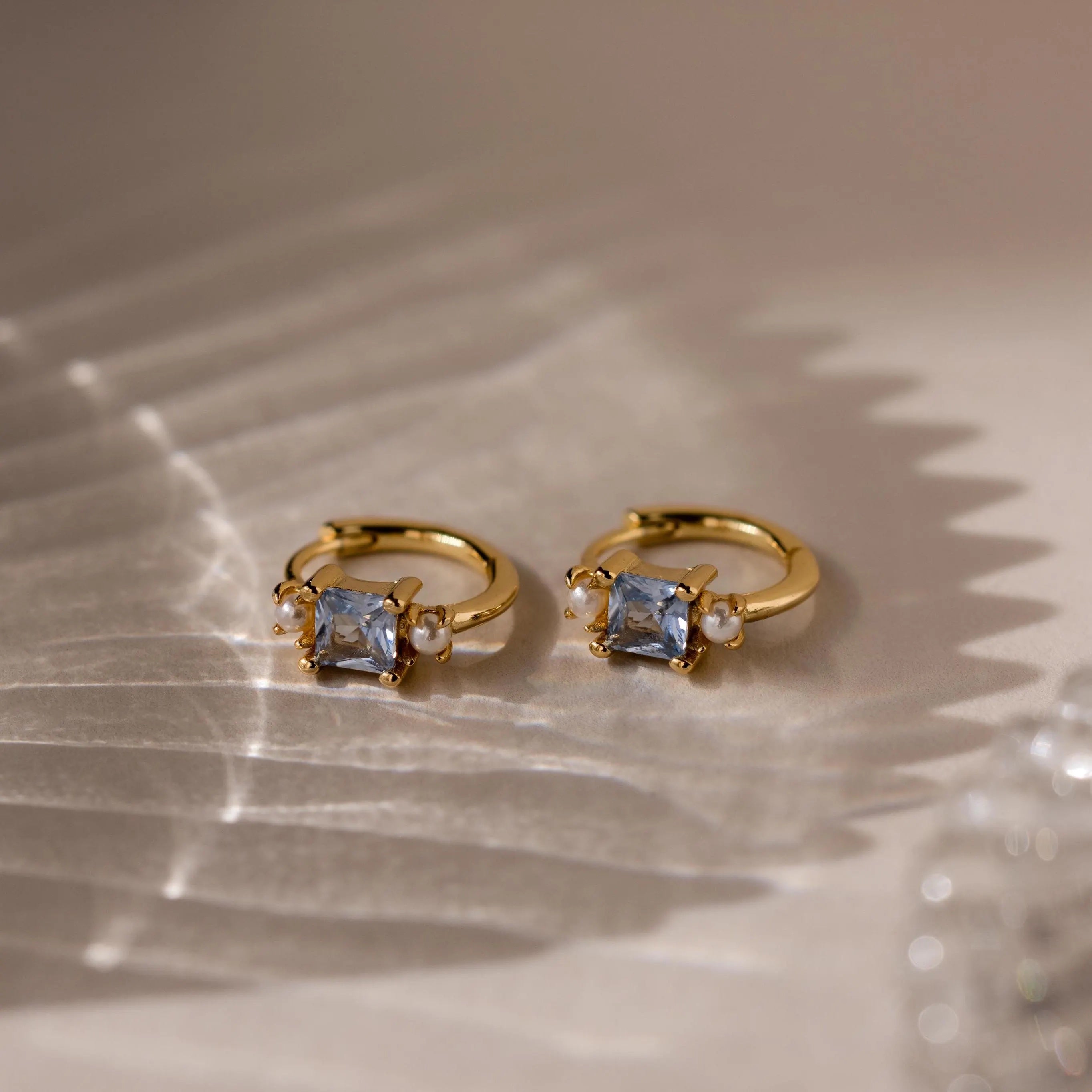 Aquamarine Pearl Huggies—gold hoop earrings with square blue gemstones and small pearls, perfect as bridal jewelry or wedding earrings—shown on a textured neutral surface.