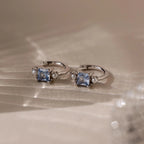 Aquamarine Pearl Huggies are silver hoop earrings featuring square blue gemstones and small pearls, making them stunning bridal jewelry or wedding earrings, beautifully displayed on a soft, light-colored surface.