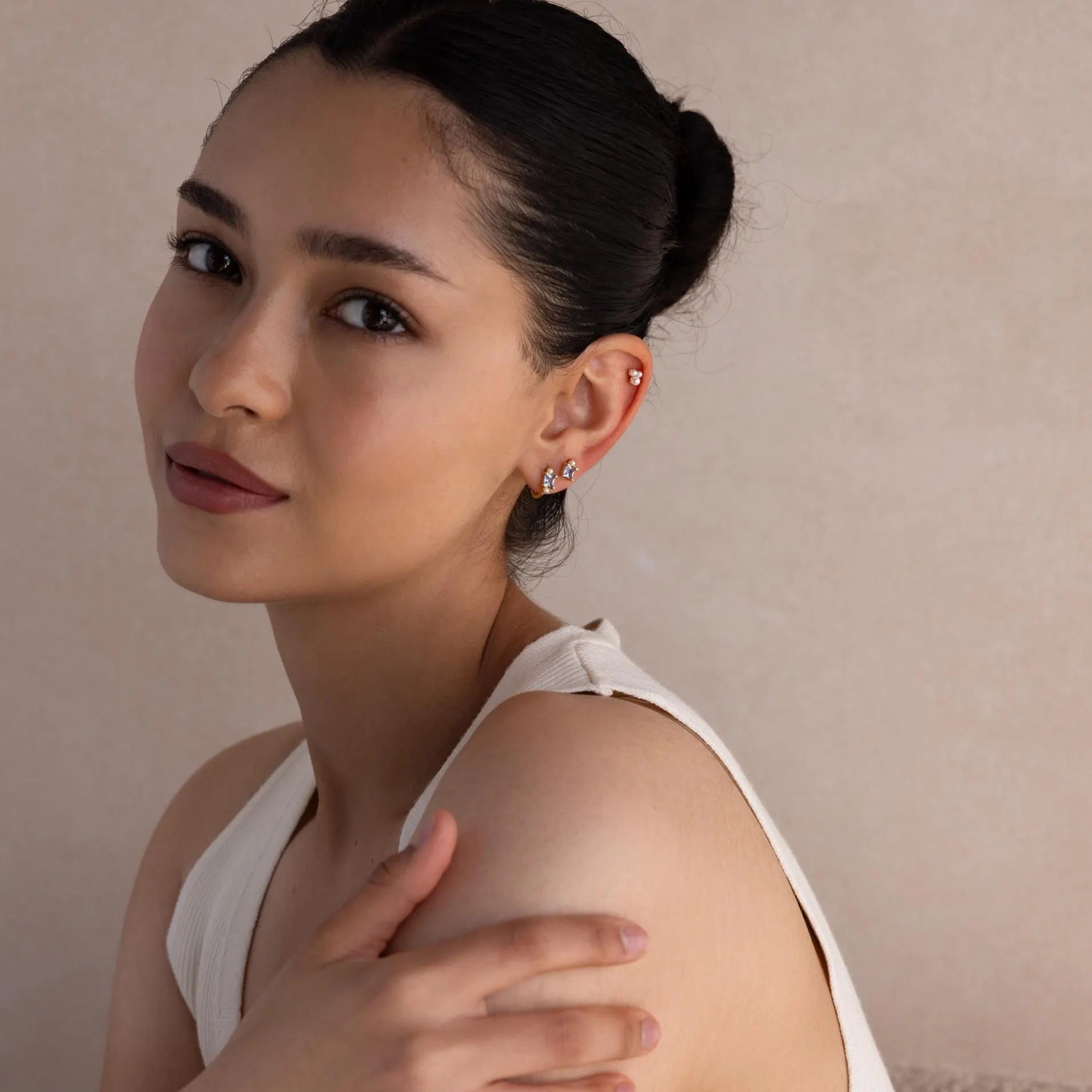 A woman with dark hair in a bun, wearing a sleeveless top and Aquamarine Pearl Huggies earrings, gazes at the camera with her hand resting on her shoulder.