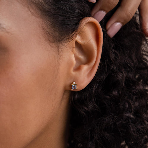Close-up of a woman's ear wearing Aquamarine Pearl Studs—small, rectangular earrings with princess-cut light blue gemstones, ideal as bridal accessories.
