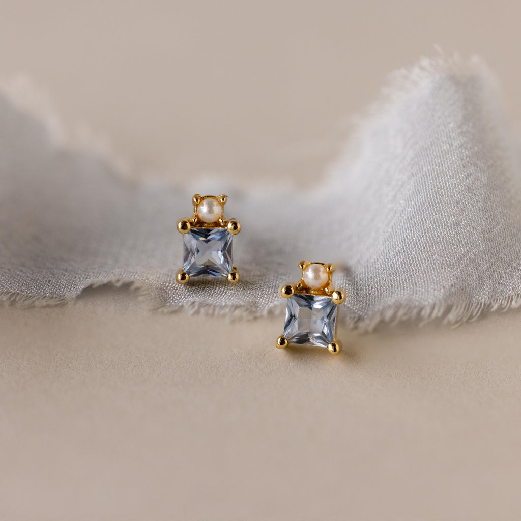 The Aquamarine Pearl Studs feature gold settings with square princess-cut aquamarines and delicate pearls, displayed on a soft, frayed fabric—perfect as elegant bridal earrings.