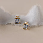 The Aquamarine Pearl Studs feature gold settings with square princess-cut aquamarines and delicate pearls, displayed on a soft, frayed fabric—perfect as elegant bridal earrings.
