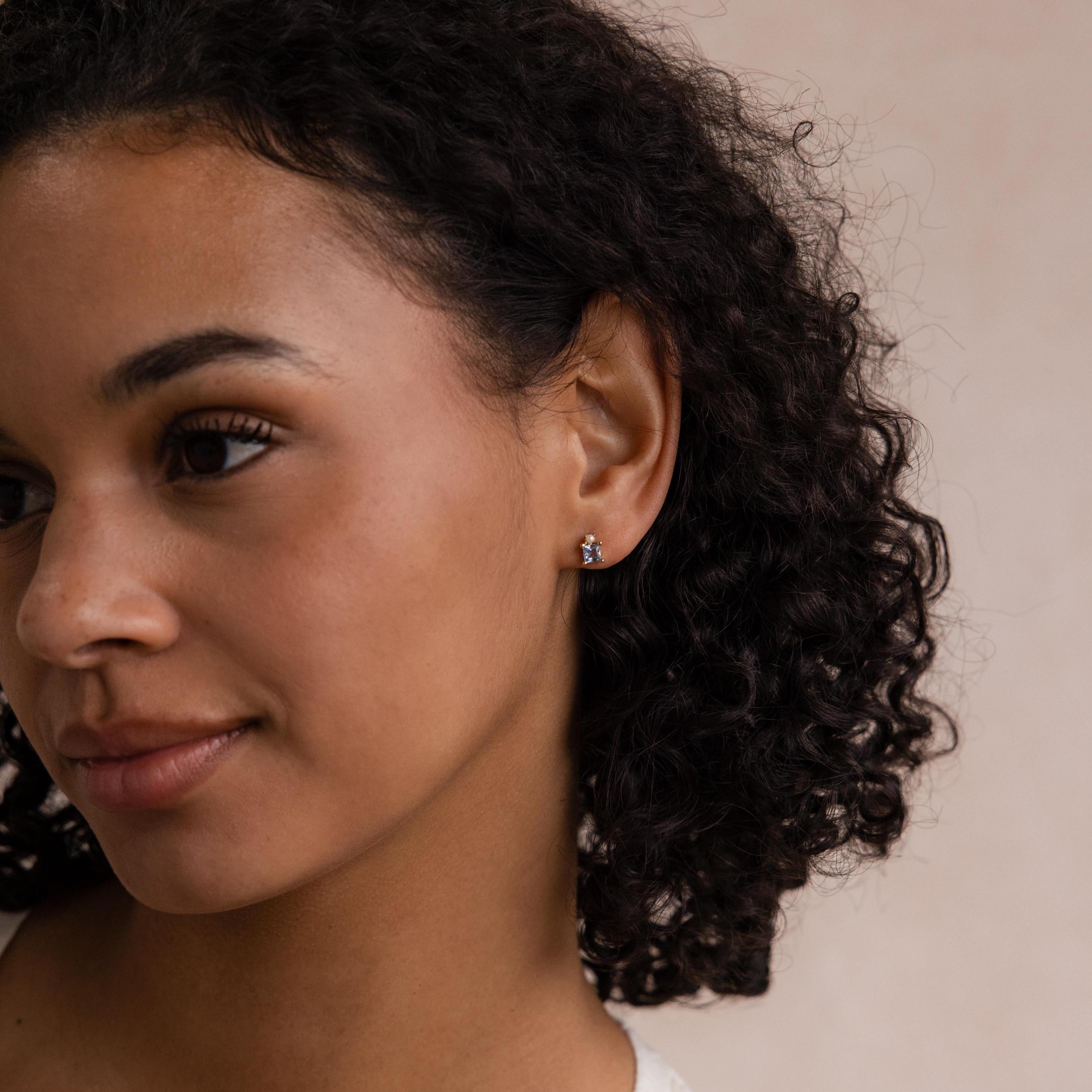 A woman with curly hair wears delicate Aquamarine Pearl Studs and smiles softly, looking to the side.