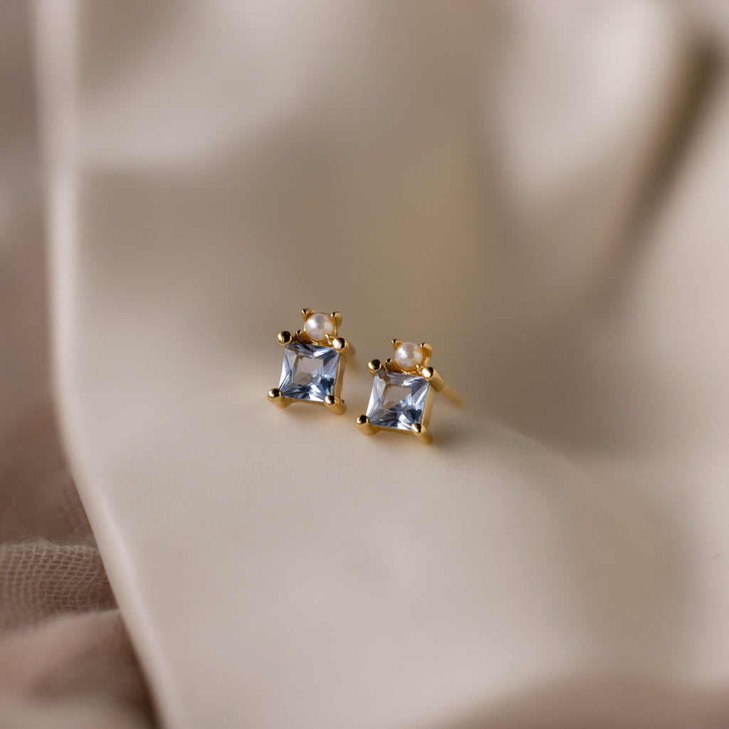 A pair of Aquamarine Pearl Studs featuring square, princess-cut gemstones is displayed on a soft beige fabric background.