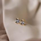 A pair of Aquamarine Pearl Studs featuring square, princess-cut gemstones is displayed on a soft beige fabric background.