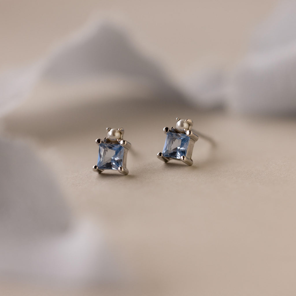 Aquamarine Pearl Studs: A pair of silver bridal stud earrings with princess-cut light blue gemstones, showcased on a soft beige background.