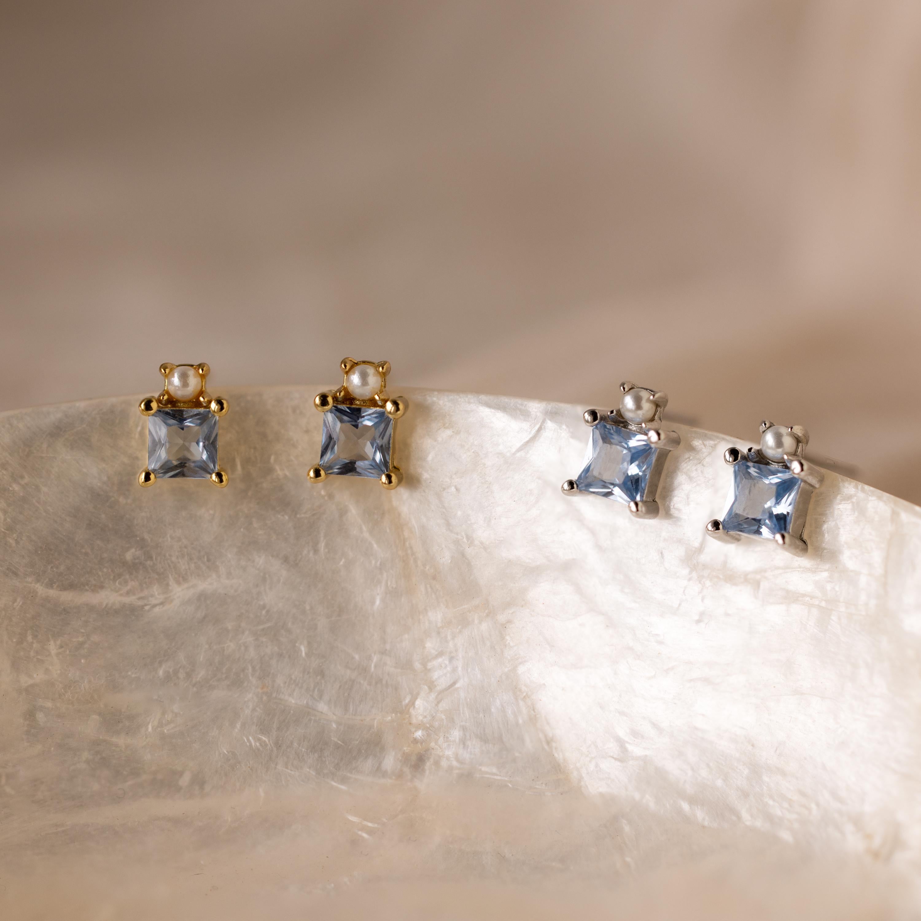 Two pairs of Aquamarine Pearl Studs, one set with gold prongs and one with silver, rest on a shell—perfect bridal earrings for a touch of blue elegance.