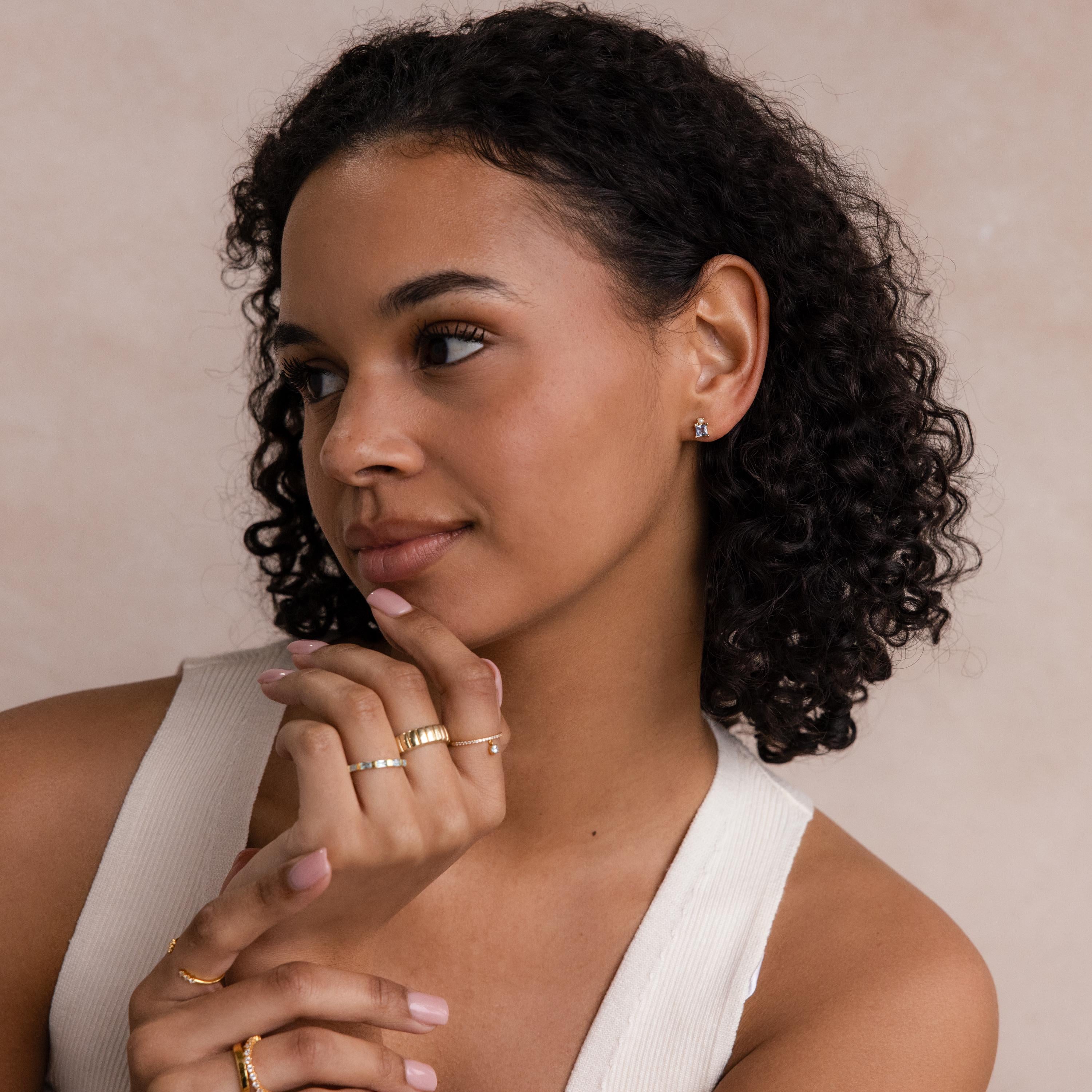 Woman with short curly hair, wearing gold rings, a sleeveless top, and Aquamarine Pearl Studs, gazes thoughtfully to the side.