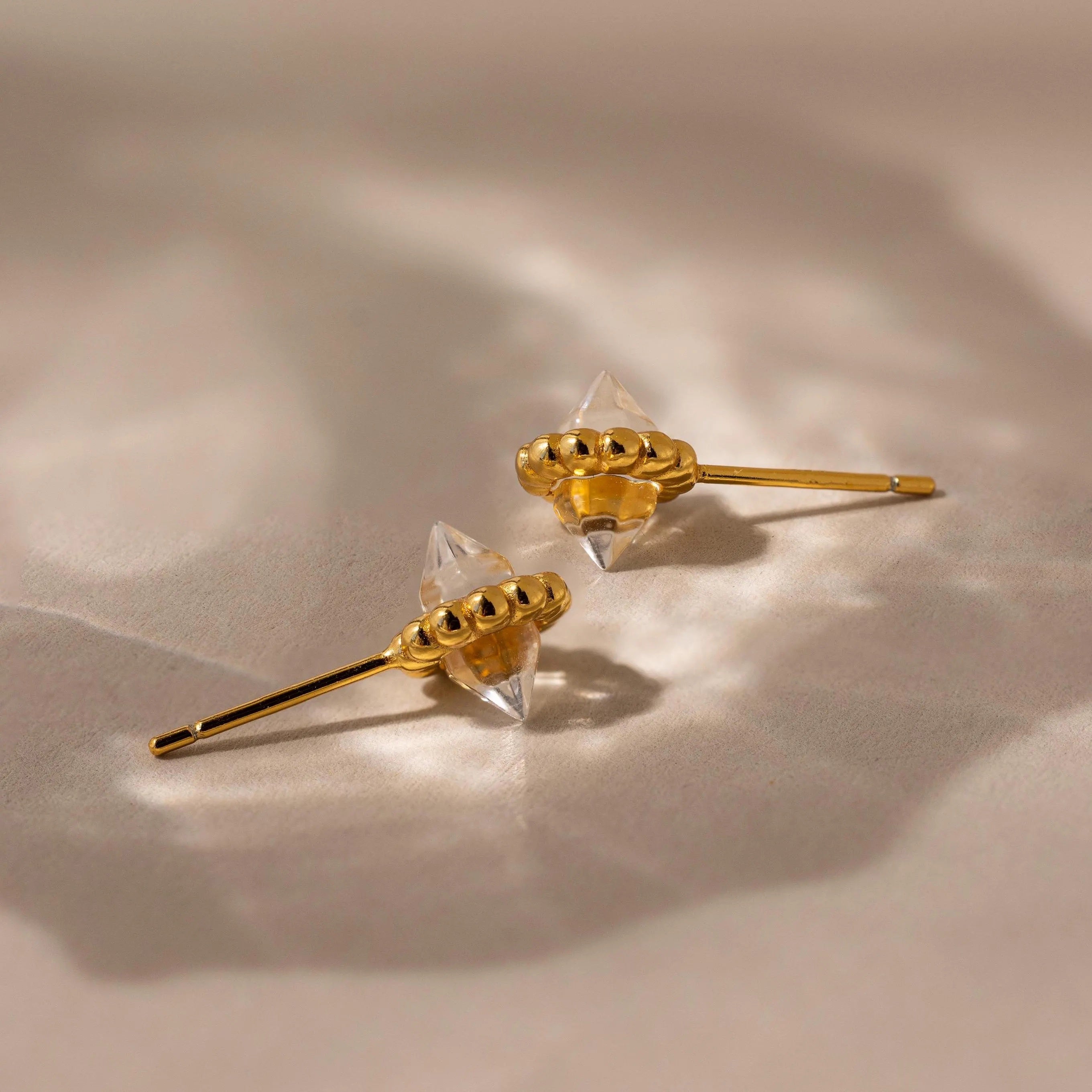 Two luminous Beaded Herkimer Studs, featuring gold and clear Herkimer stones, rest on a soft beige surface with gentle shadows.