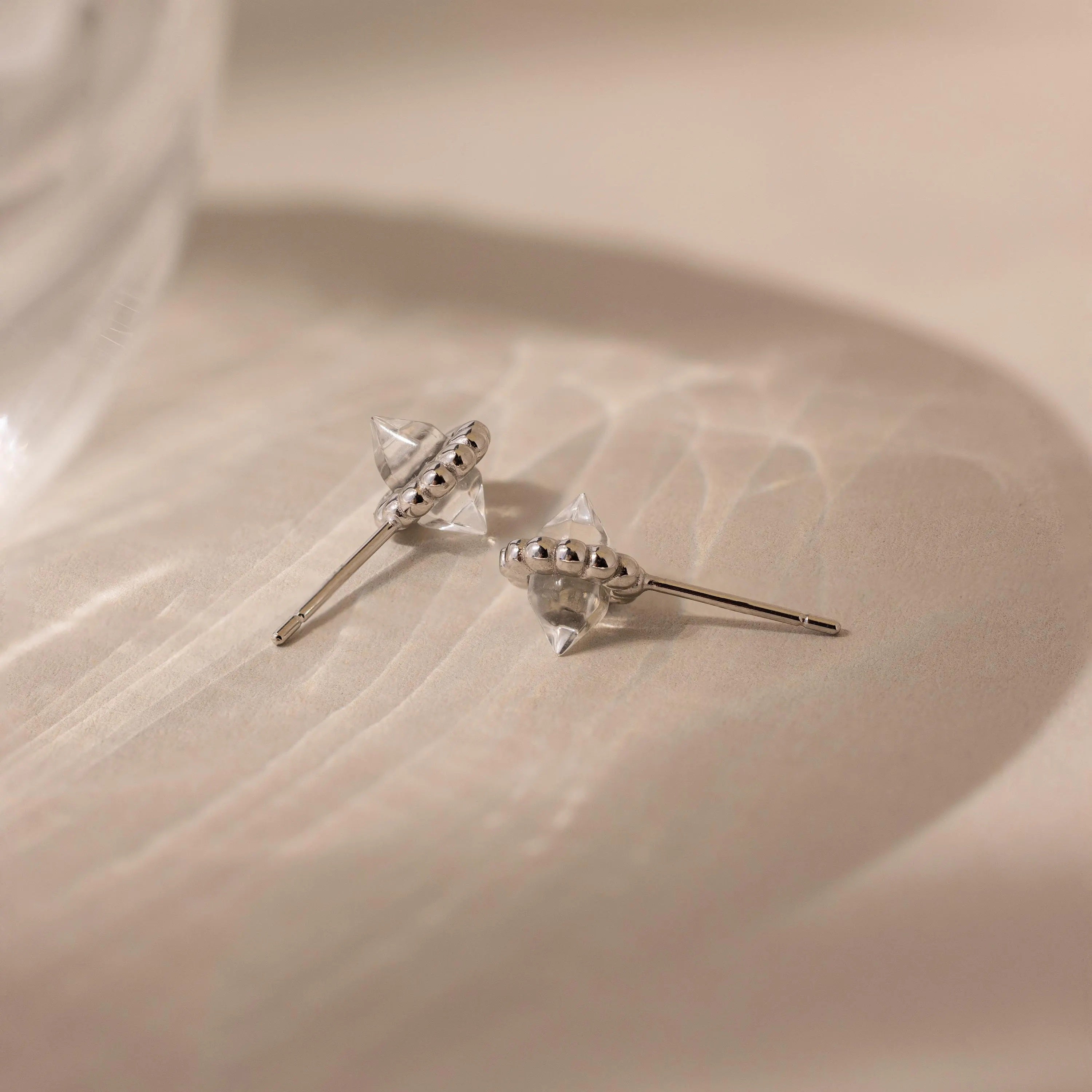 The Beaded Herkimer Studs, featuring clear faceted accents, rest on a softly textured beige surface, giving these silver earrings a luminous shine.