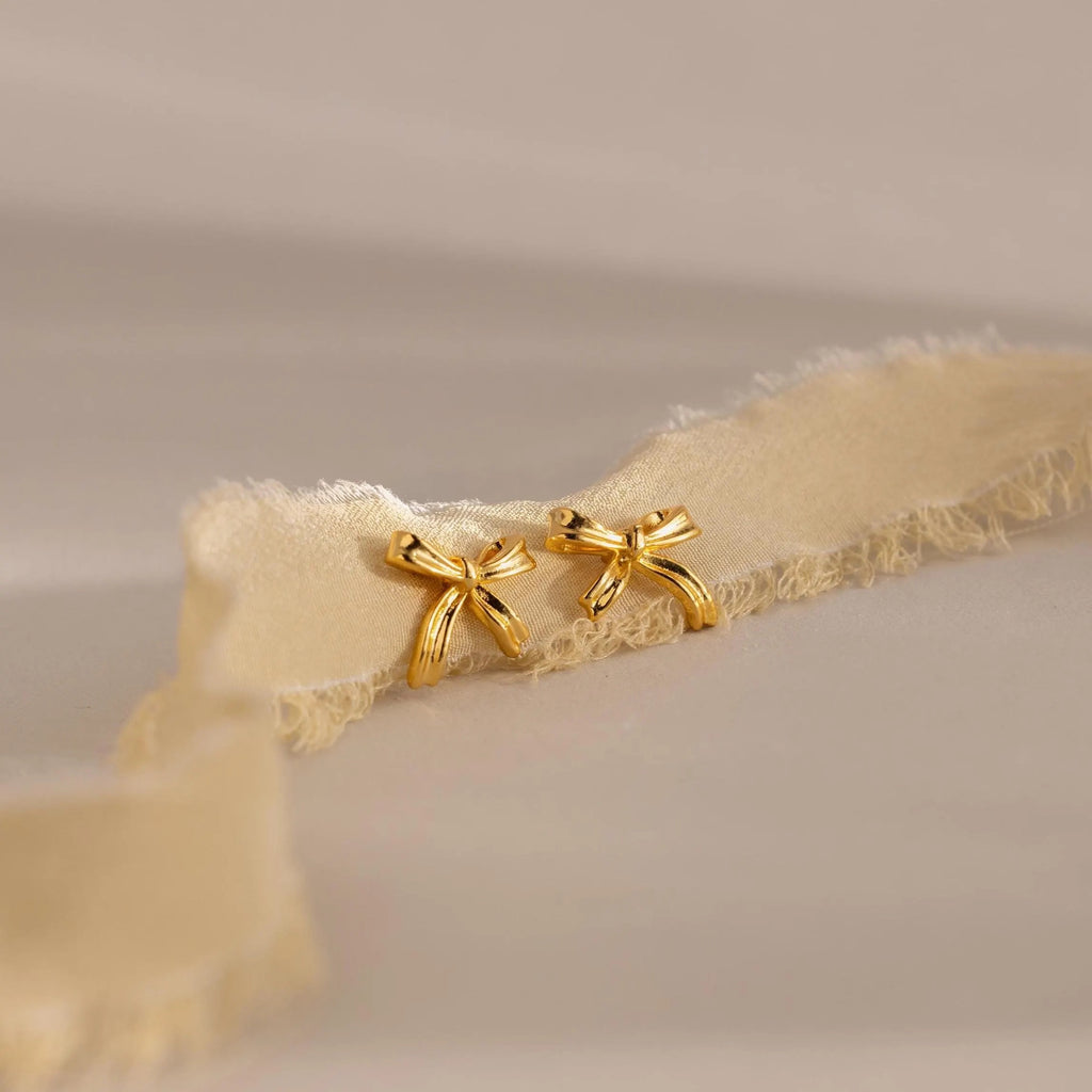 A pair of gold bow stud earrings displayed on a soft ivory ribbon background, showing their polished surface and delicate ribbon design.