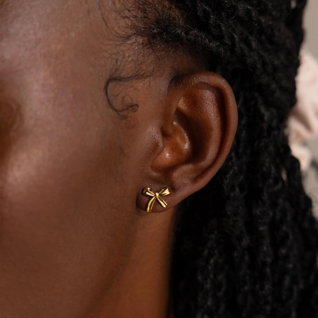 A close-up side profile of a woman wearing a single gold bow stud earring in her earlobe. The earring is delicately crafted into the shape of a petite bow with smooth, polished ribbon loops and gently curved tails, capturing the soft, feminine elegance of a tied ribbon.