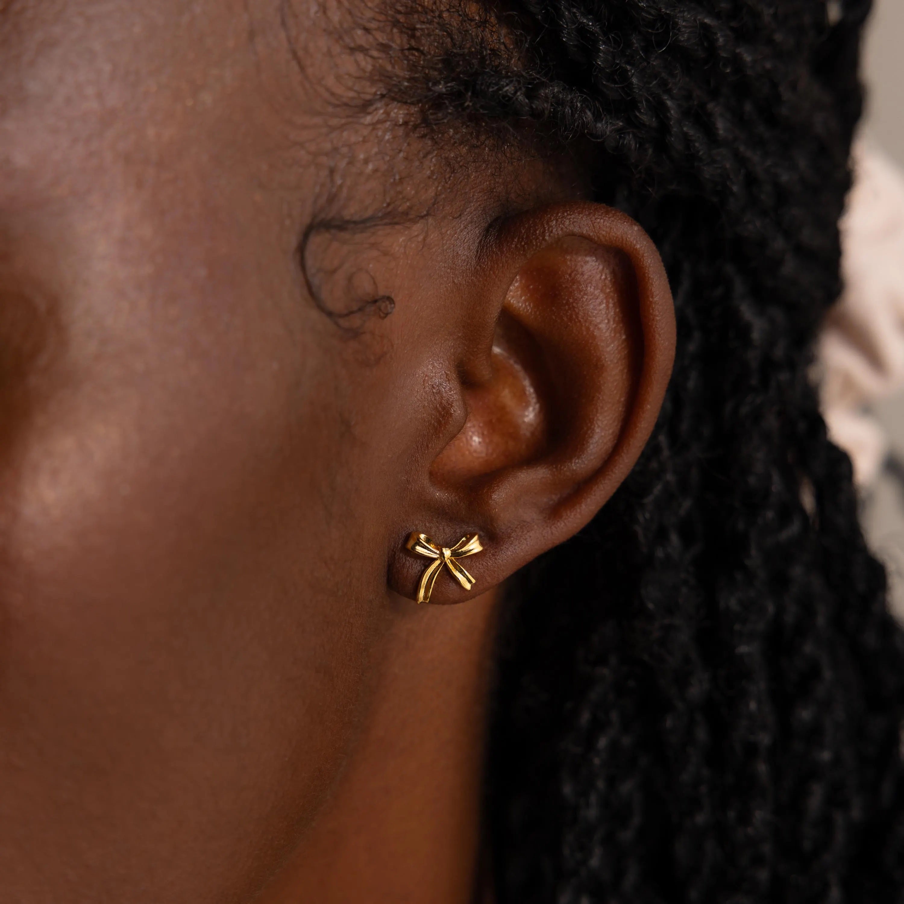 A close-up side profile of a woman wearing a single gold bow stud earring in her earlobe. The earring is delicately crafted into the shape of a petite bow with smooth, polished ribbon loops and gently curved tails, capturing the soft, feminine elegance of a tied ribbon.