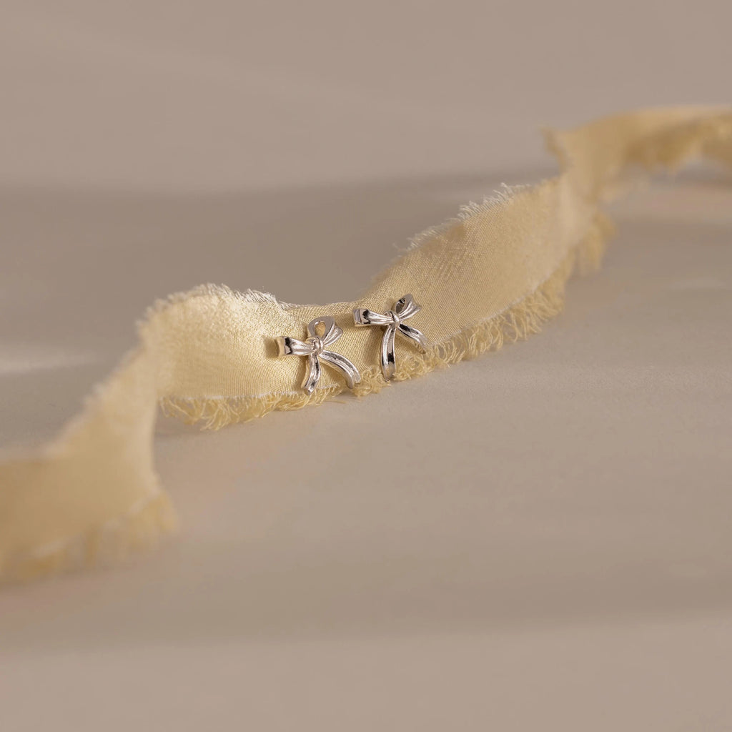 A pair of silver bow stud earrings placed on an ivory frayed-edge fabric.