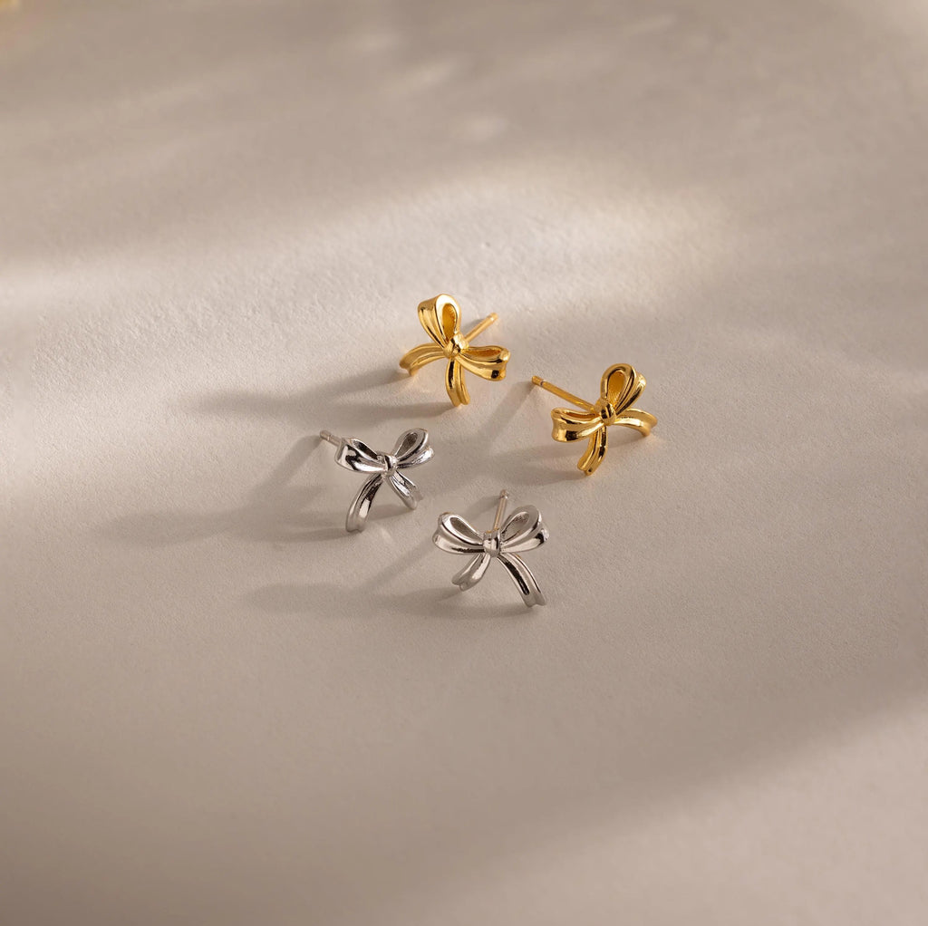 Product flat lay featuring two pairs of bow stud earrings—one gold and one white gold—arranged under soft lighting on a neutral background.