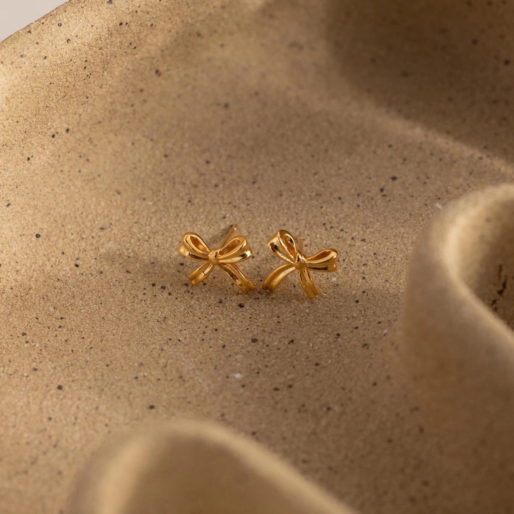 Gold bow stud earrings resting on a bed of sand, accentuating their warm tone and soft, graceful curves.