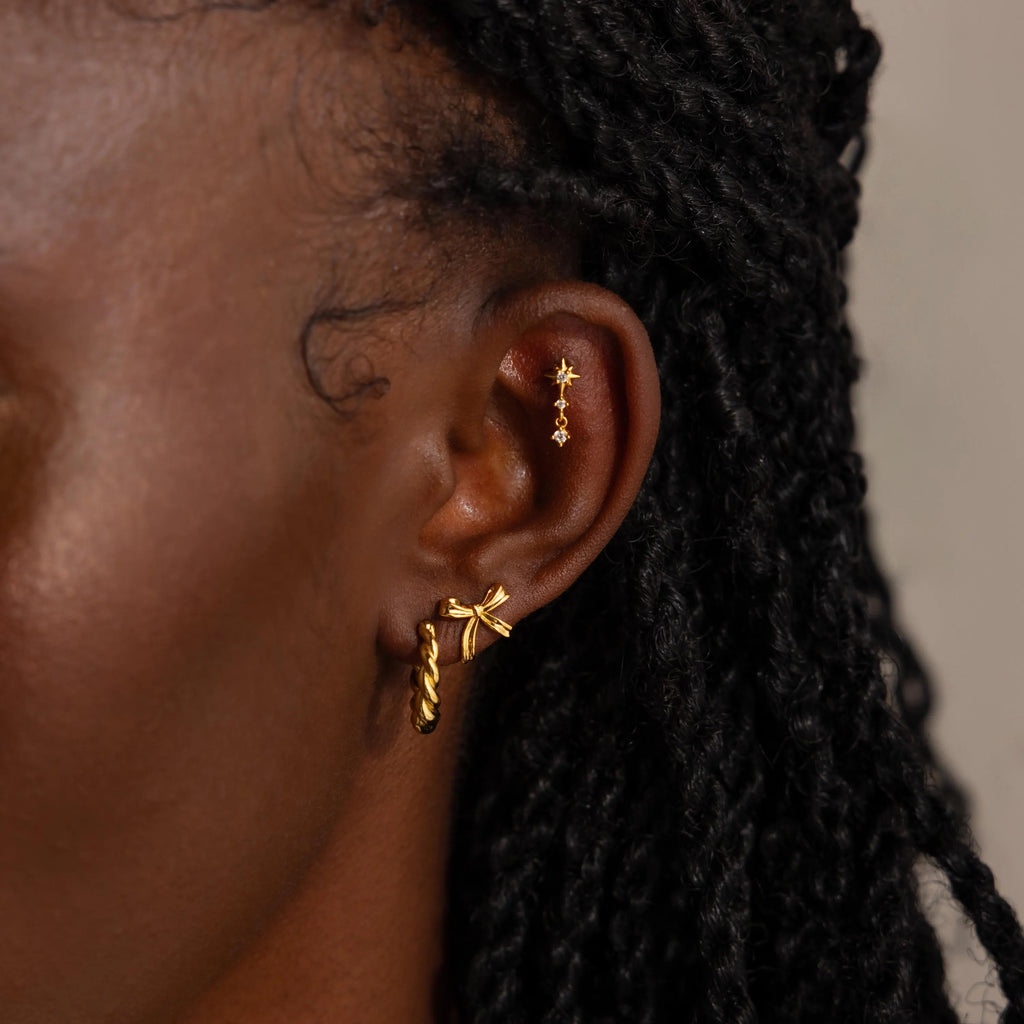 Portrait of the woman styled with multiple earrings in her ear, including the gold bow studs on her lobe, layered with other gold jewelry along her helix and upper ear.