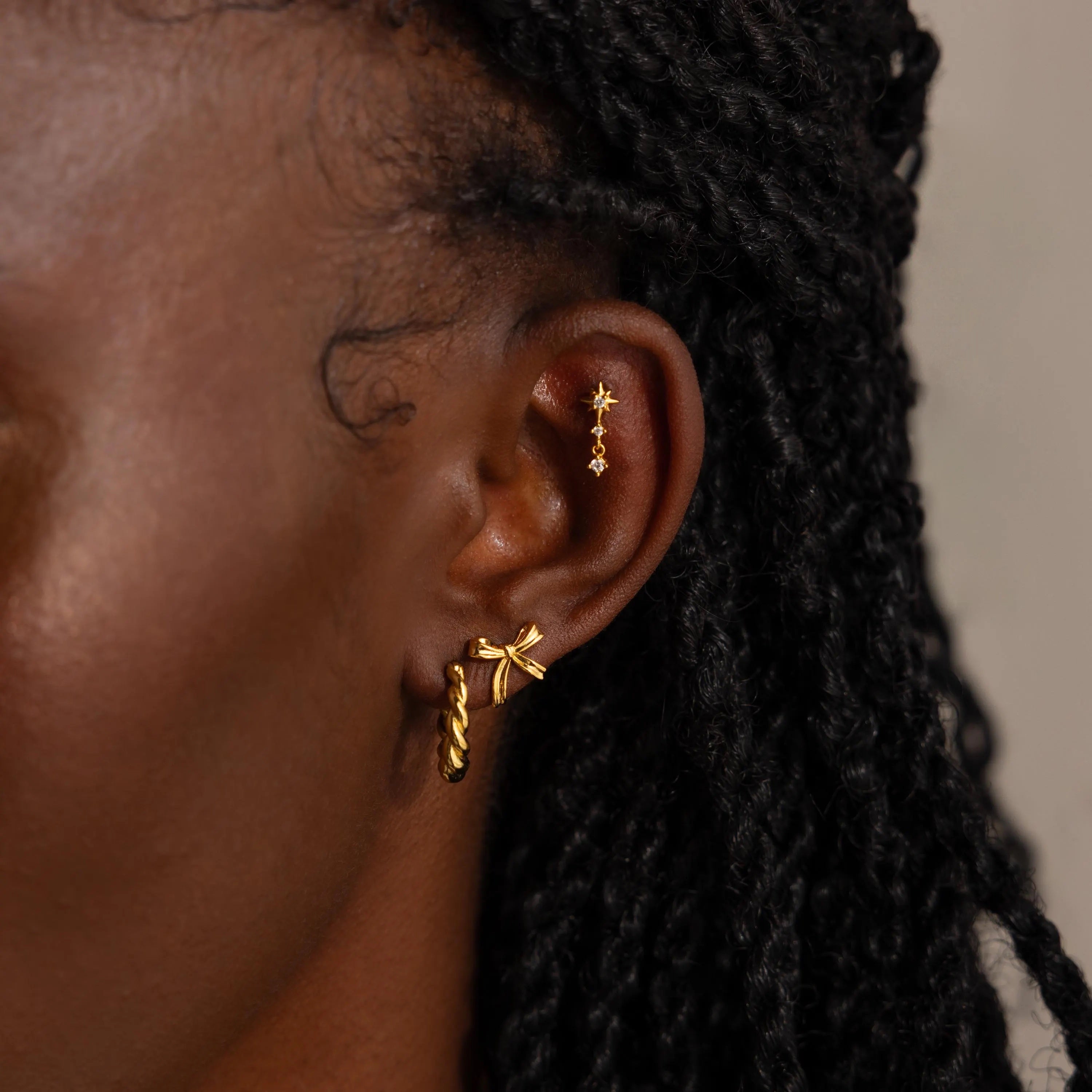Portrait of the woman styled with multiple earrings in her ear, including the gold bow studs on her lobe, layered with other gold jewelry along her helix and upper ear.