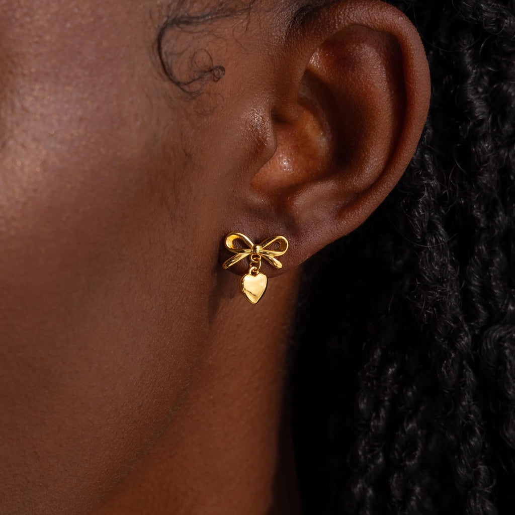 A close-up view of a woman’s ear adorned with a delicate gold stud earring designed in the shape of a dainty bow. Dangling just below the bow is a small, highly polished heart charm that gently catches the light.