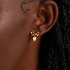 A close-up view of a woman’s ear adorned with a delicate gold stud earring designed in the shape of a dainty bow. Dangling just below the bow is a small, highly polished heart charm that gently catches the light.