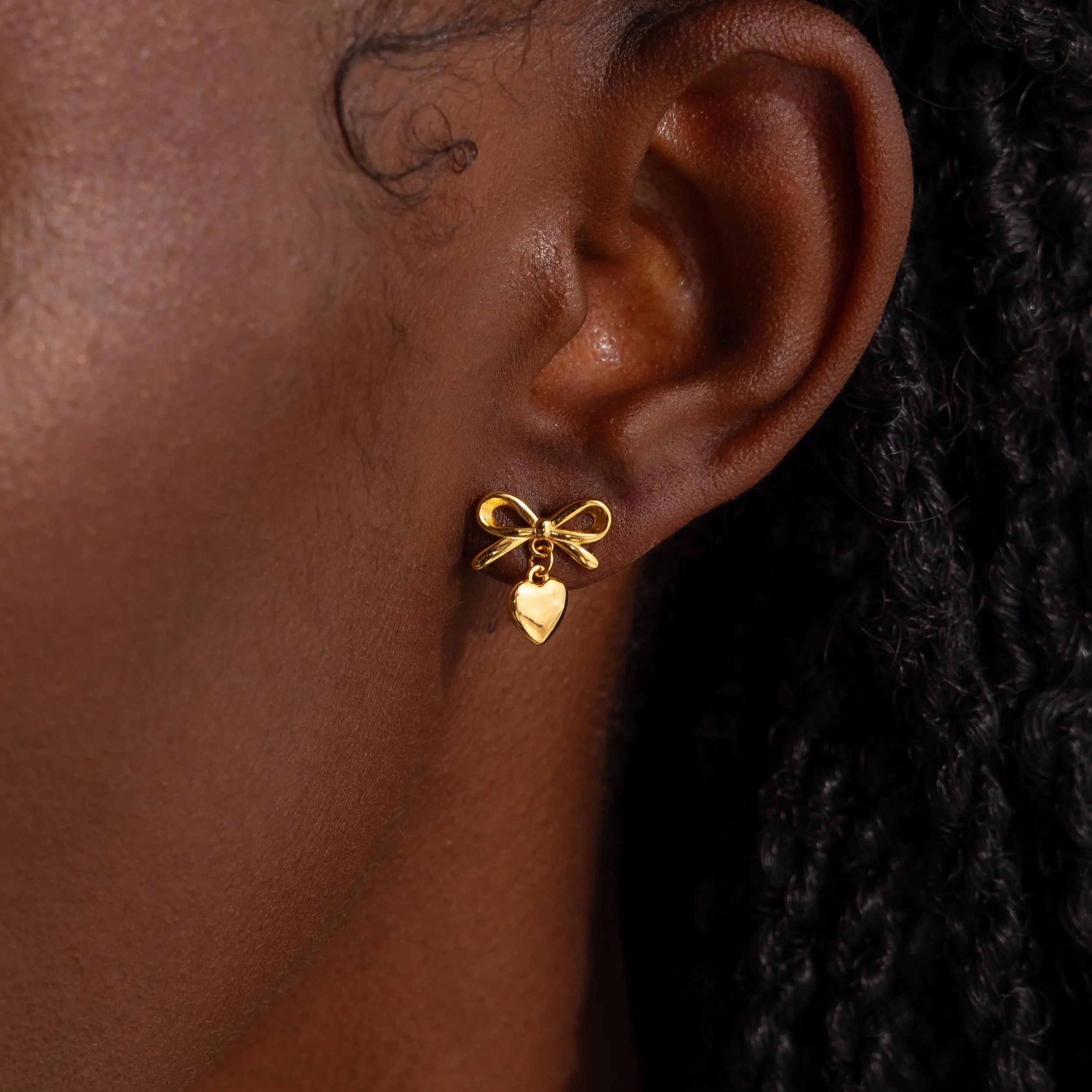 A close-up view of a woman’s ear adorned with a delicate gold stud earring designed in the shape of a dainty bow. Dangling just below the bow is a small, highly polished heart charm that gently catches the light.