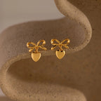 A pair of gold bow-shaped stud earrings with heart charms displayed on a sculptural, neutral-toned ceramic surface.