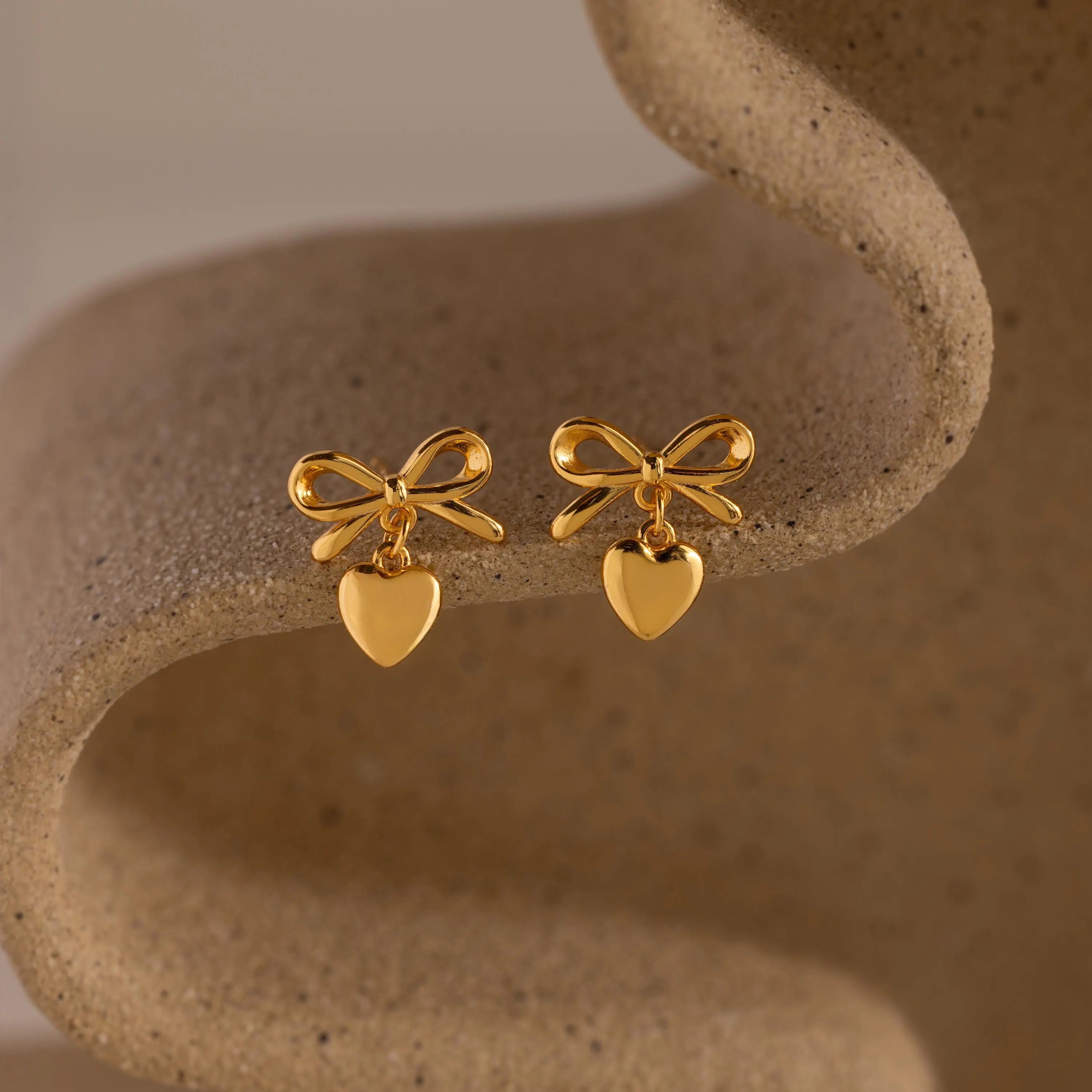 A pair of gold bow-shaped stud earrings with heart charms displayed on a sculptural, neutral-toned ceramic surface.