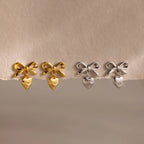 Two pairs of bow heart stud earrings displayed on a torn-edge neutral backdrop—one pair in gold, the other in silver.