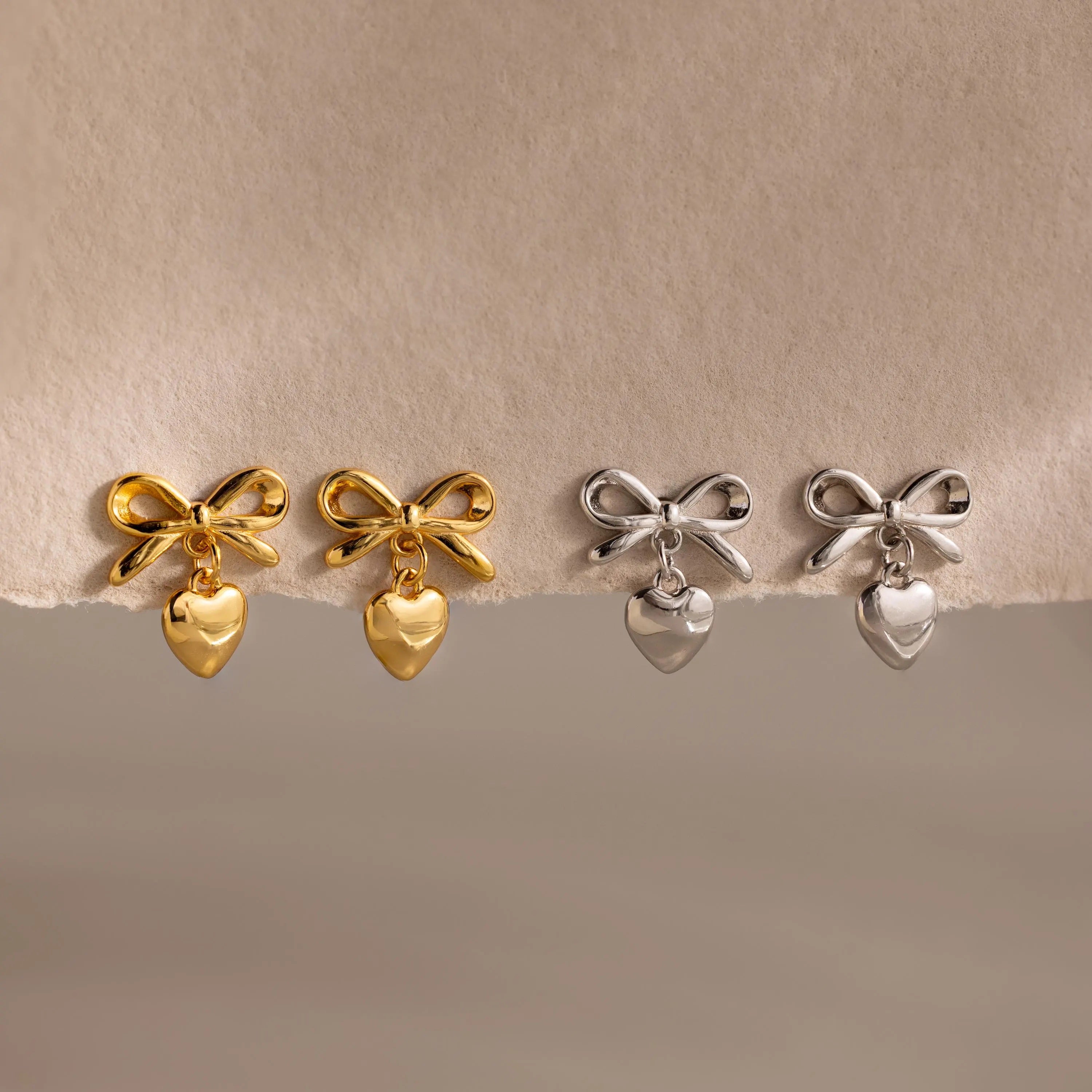 Two pairs of bow heart stud earrings displayed on a torn-edge neutral backdrop—one pair in gold, the other in silver.