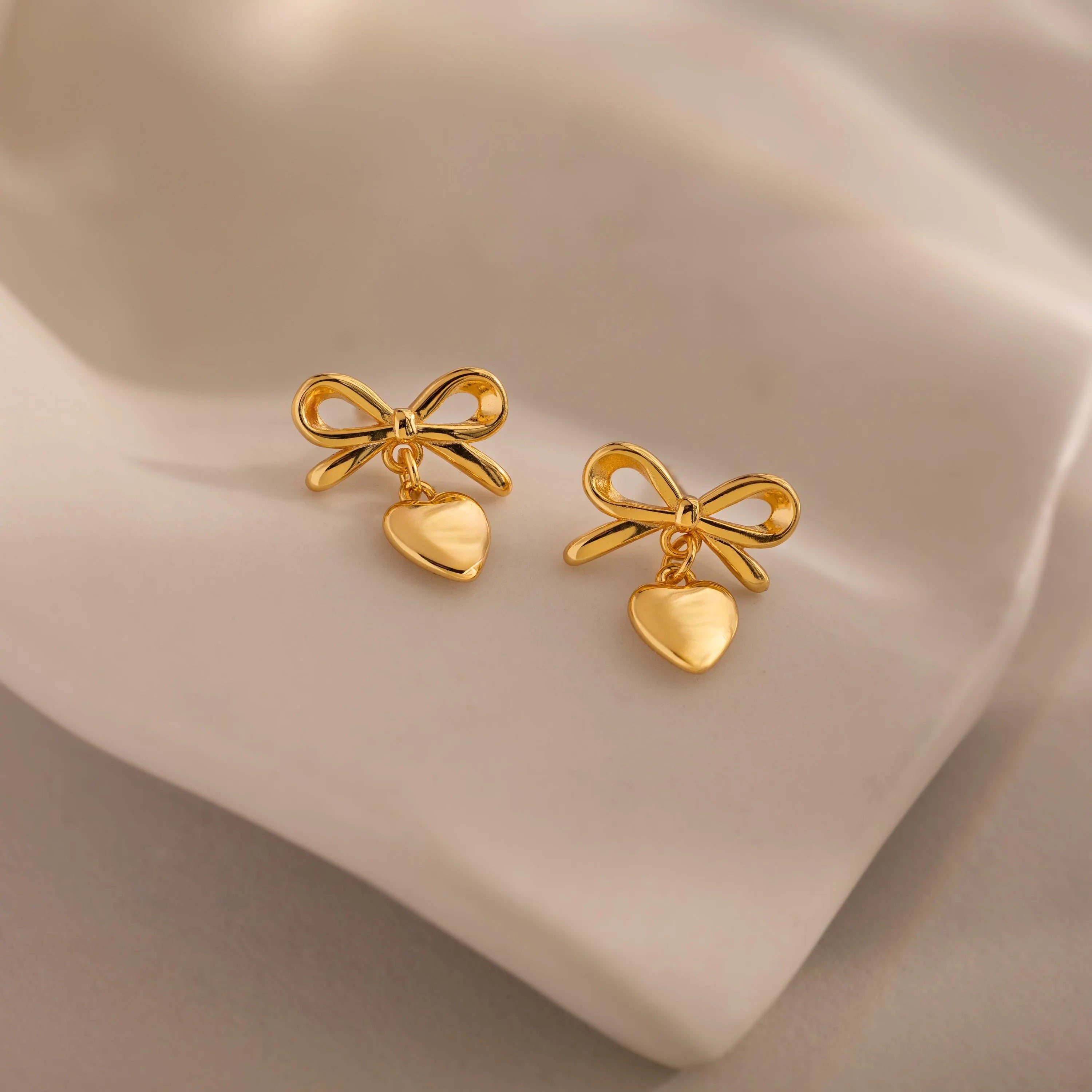 A pair of gold bow heart stud earrings resting on a smooth ivory surface, capturing the sheen of the polished finish.