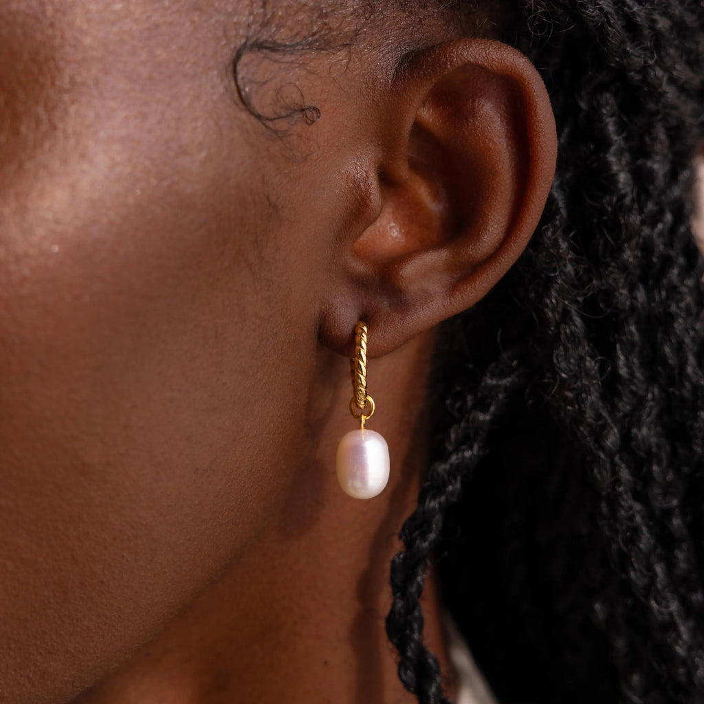 A close-up of an ear adorned with Twisted Pearl Hoops—gold spiral hoops featuring a dangling white pearl that exude elegance.
