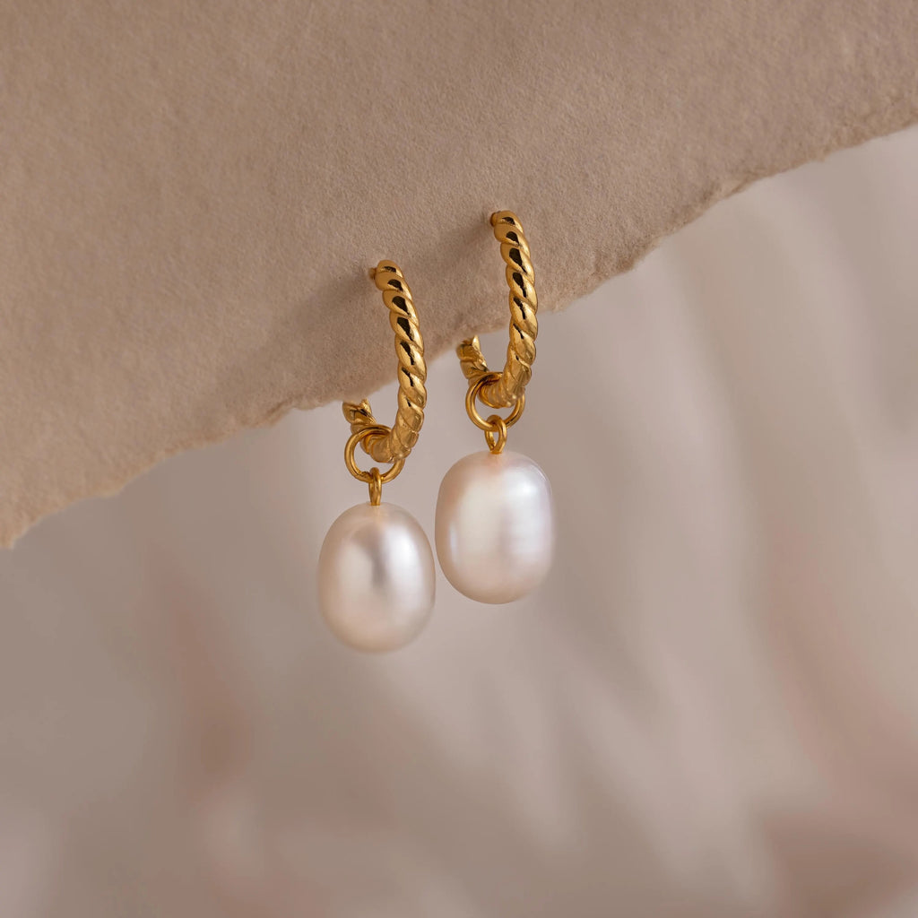 Pair of gold twisted hoop earrings each featuring a single dangling white freshwater pearl, displayed against a soft neutral background.