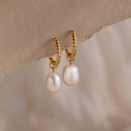Pair of gold twisted hoop earrings each featuring a single dangling white freshwater pearl, displayed against a soft neutral background.