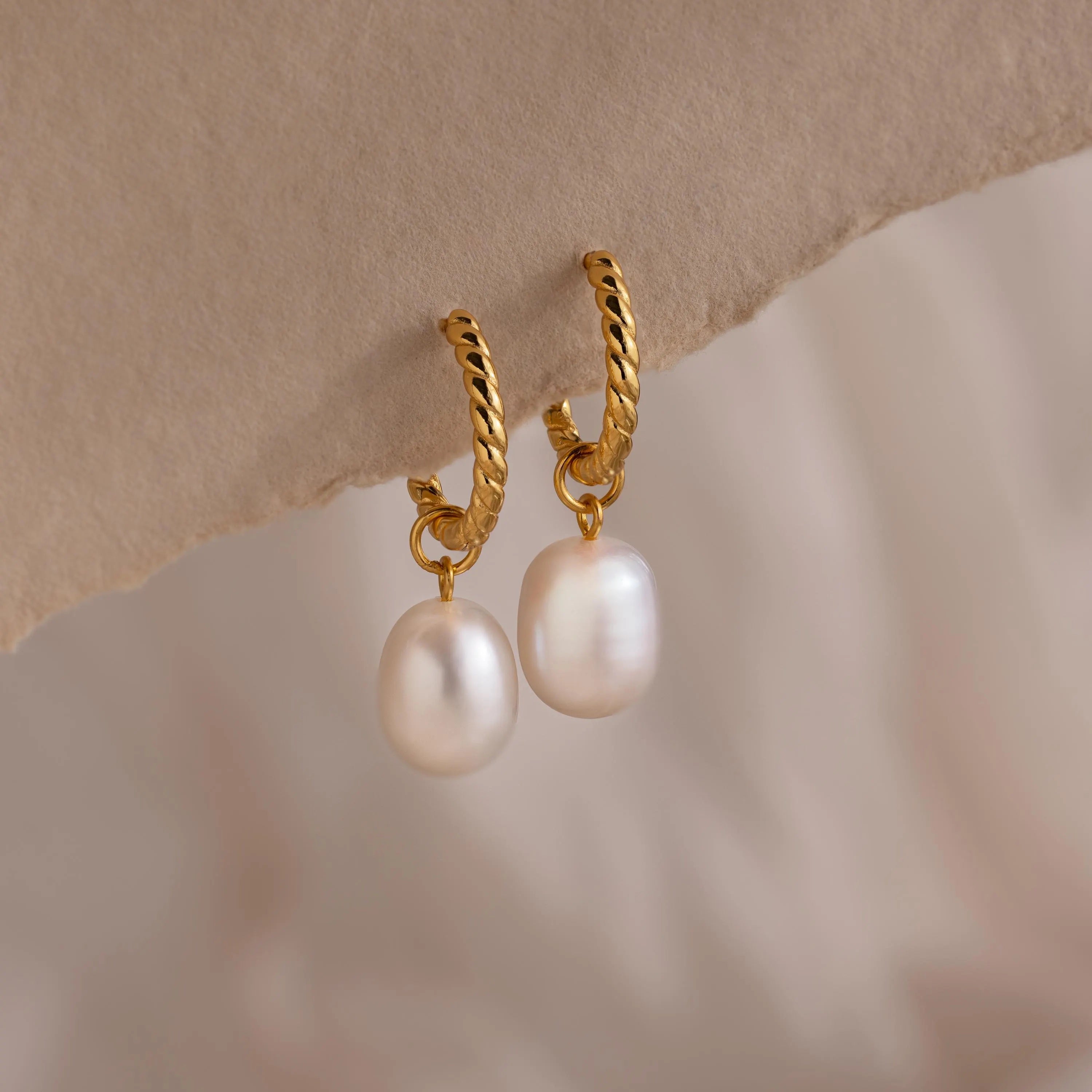Pair of gold twisted hoop earrings each featuring a single dangling white freshwater pearl, displayed against a soft neutral background.