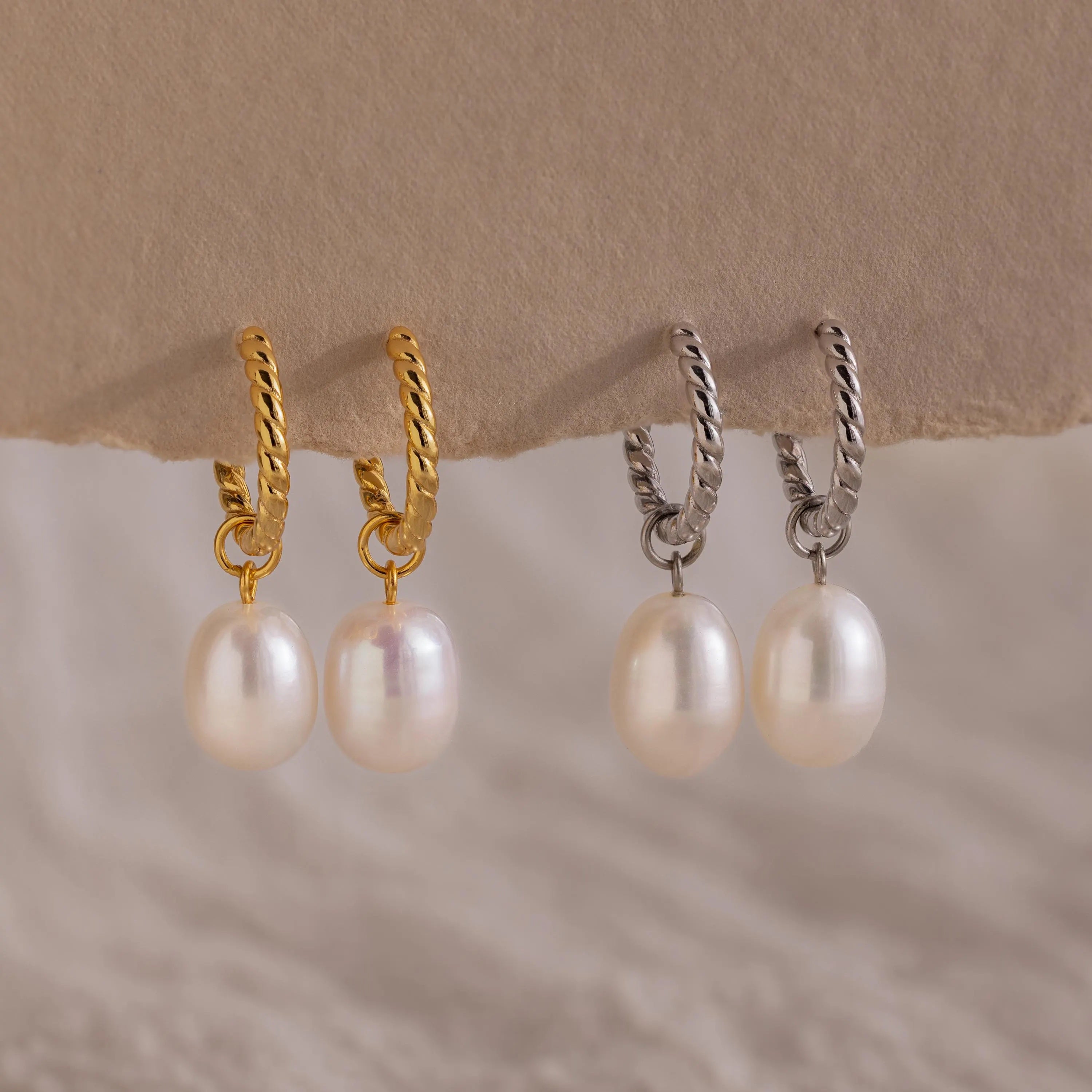 Side-by-side comparison of the gold and silver versions of the Twisted Pearl Hoop Earrings, all four displayed hanging from a neutral paper edge.