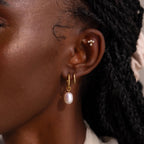 Close-up of a woman’s ear with a layered earring look, wearing the Twisted Pearl Hoop Earring in gold alongside a plain gold hoop and a tiny pearl ear cuff.