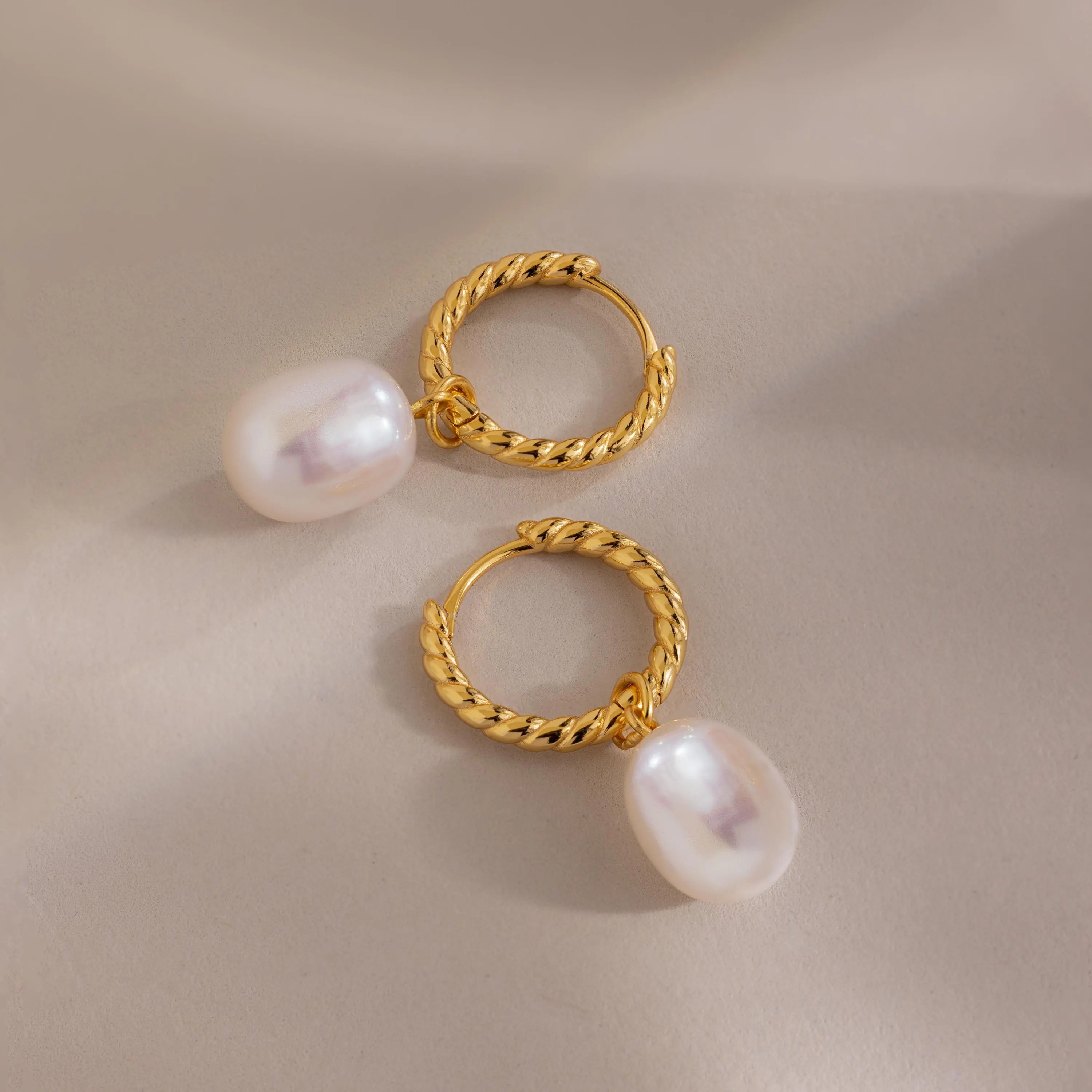 Two gold twisted hoop earrings with pearls shown flat on a beige surface, one positioned upright and the other tilted for depth.