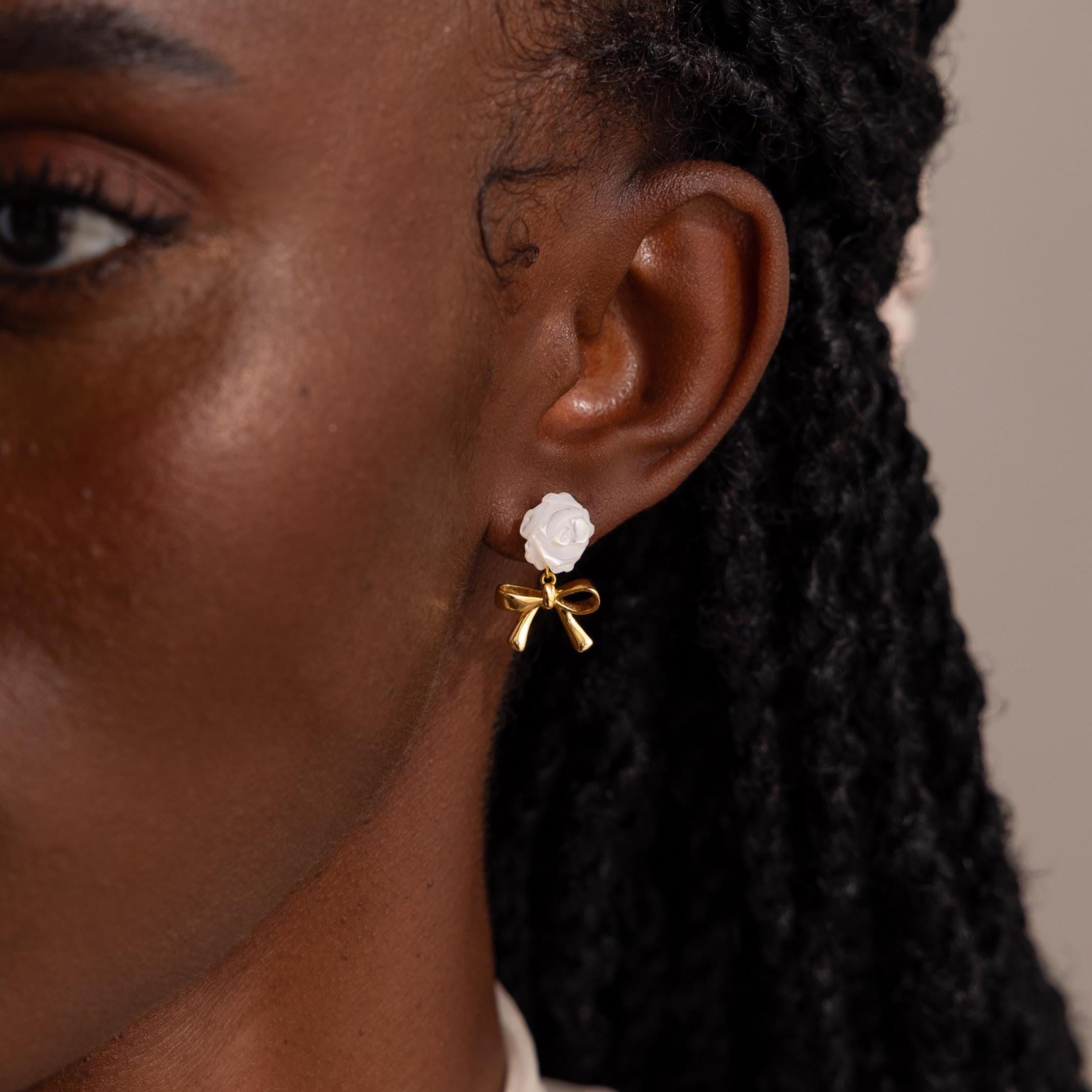 Close-up view of a woman's ear adorned with a delicate earring featuring a sculpted white rose stud and a dangling gold bow charm.