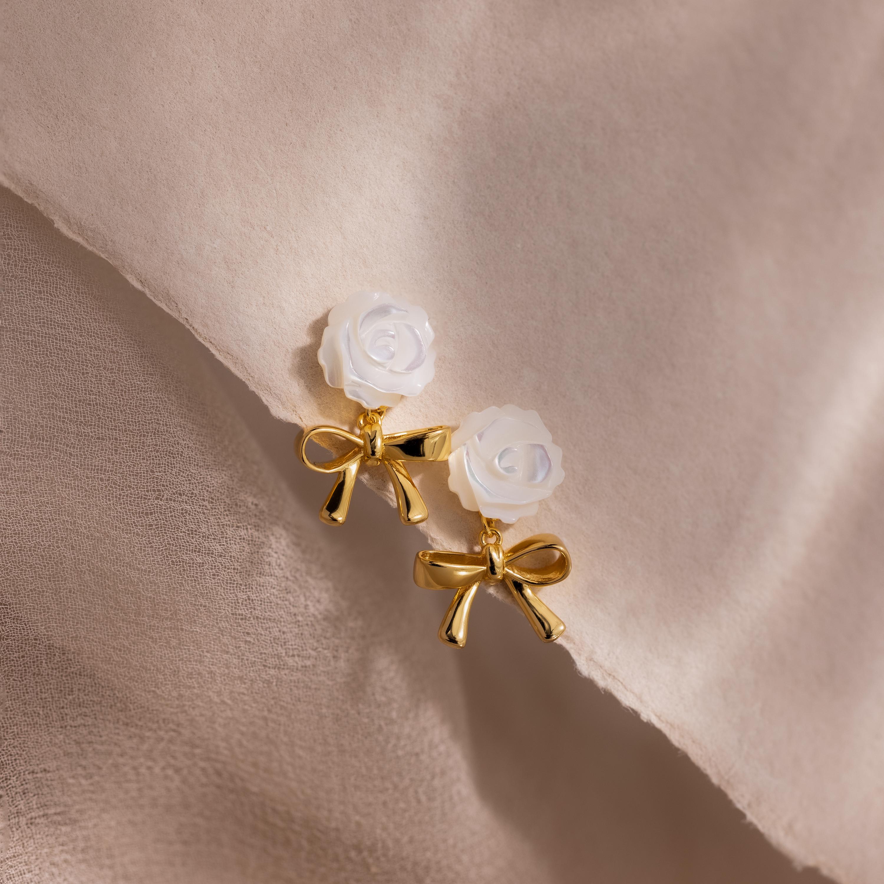 A pair of earrings with white rose studs and gold bow dangles, laid on soft neutral fabric.