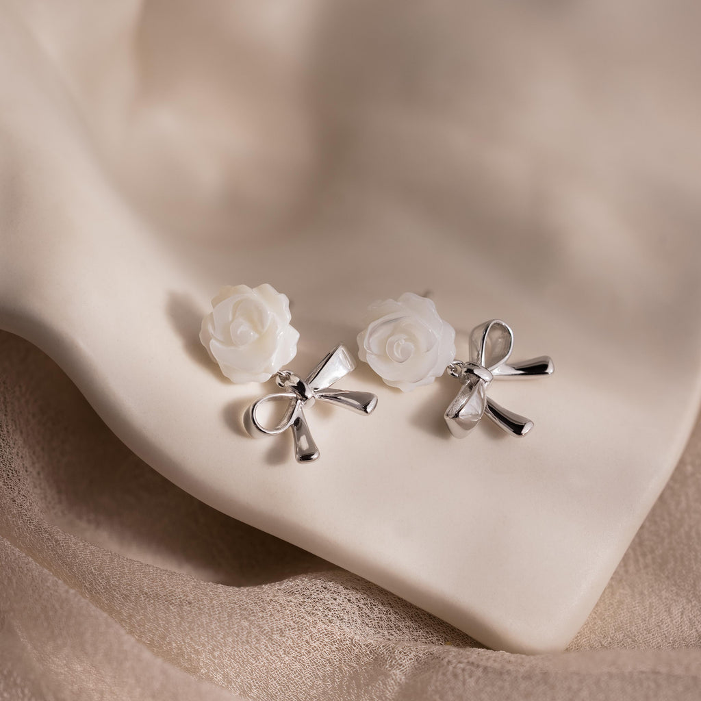 White rose and silver bow earrings displayed on light cream satin fabric.